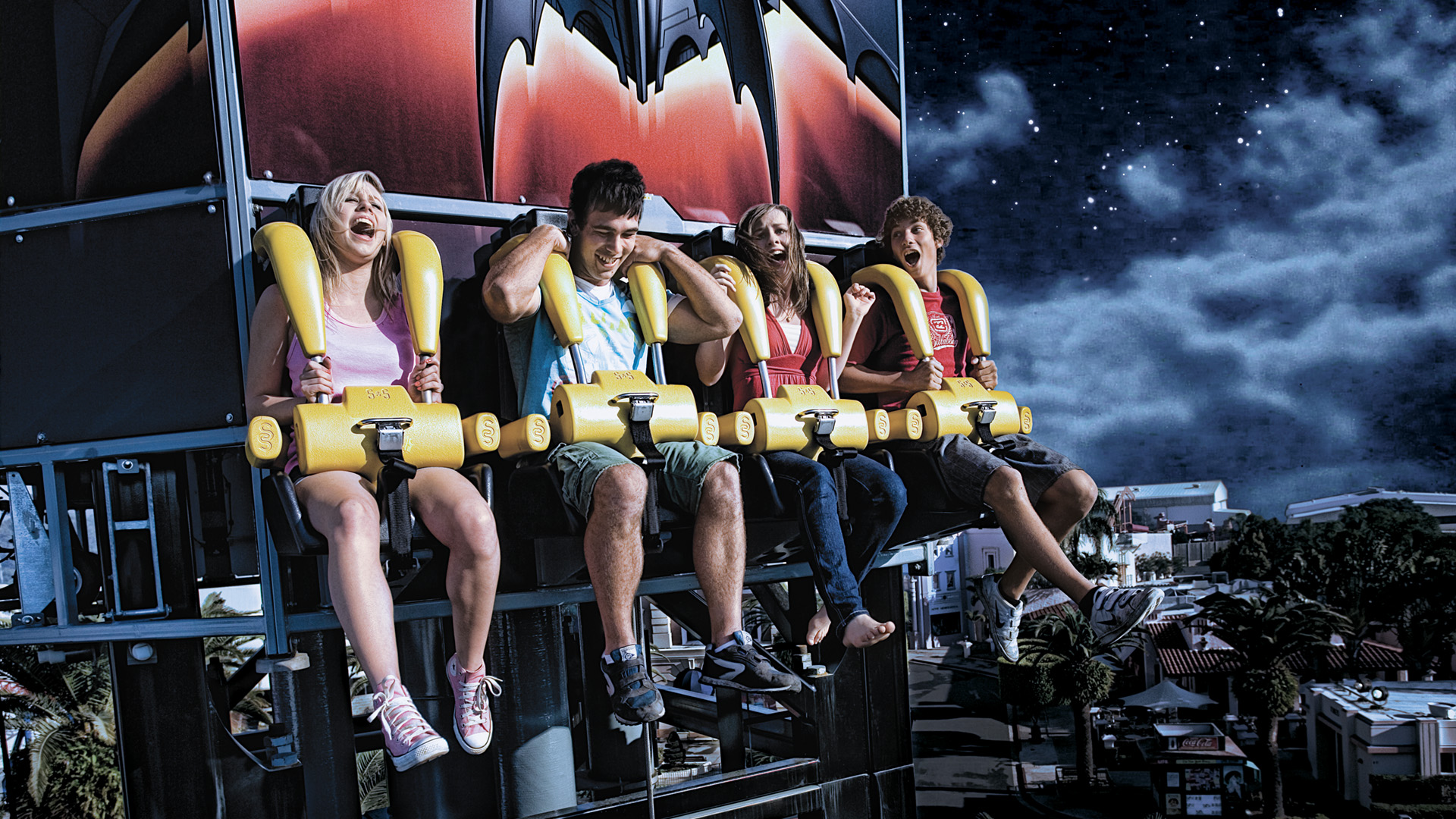 Four people are strapped into seats on a drop tower amusement park ride at night, looking excited and anxious as they prepare for the ride to begin. The background shows a starry sky and park lights below.