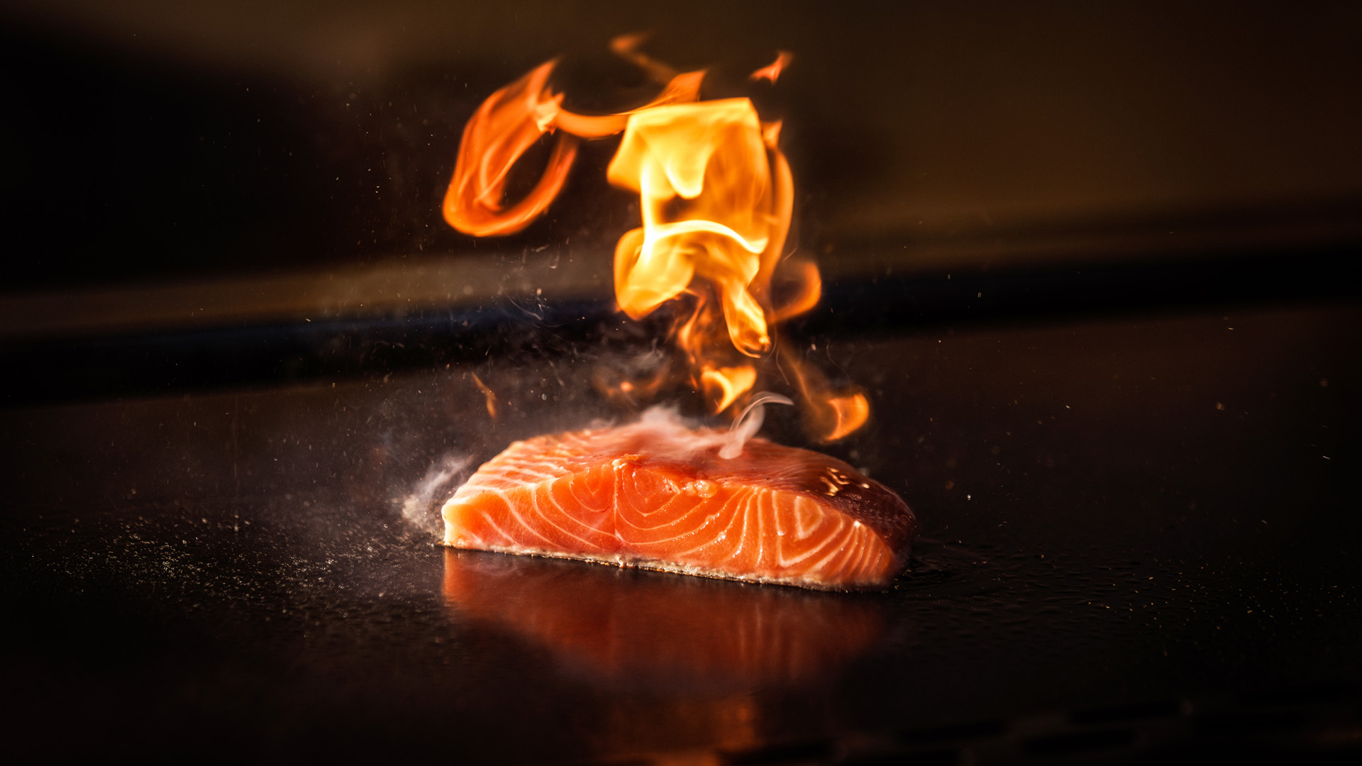 A piece of salmon is being seared on a grill, with bright orange flames rising above it, highlighting the fish's fresh, marbled texture against a dark background.