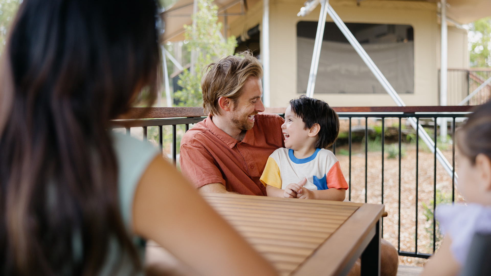 A man and a young boy sit at an outdoor table, smiling at each other, with two other people partially visible in the foreground.