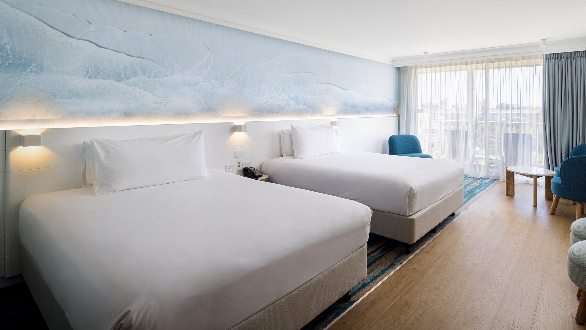 A modern hotel room with two large beds, a blue accent wall, floor-to-ceiling windows with sheer curtains, a small round table, a blue chair, and wooden floors. Natural light fills the spacious room.
