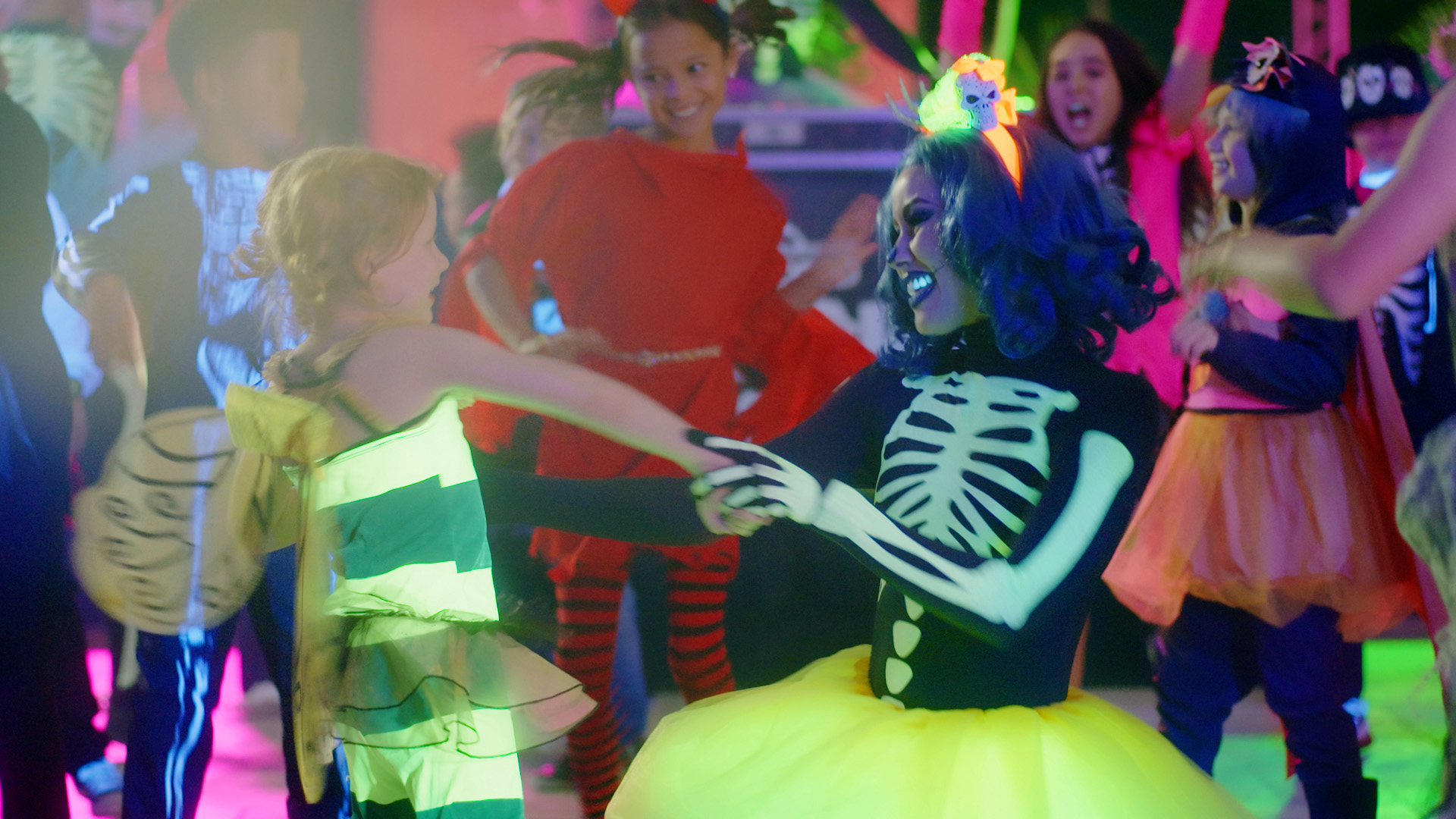 Children in colorful costumes dance at a party. One child is dressed as a bee and another as a skeleton with a yellow tutu and a bright wig. Other kids in festive costumes are visible in the lively background.
