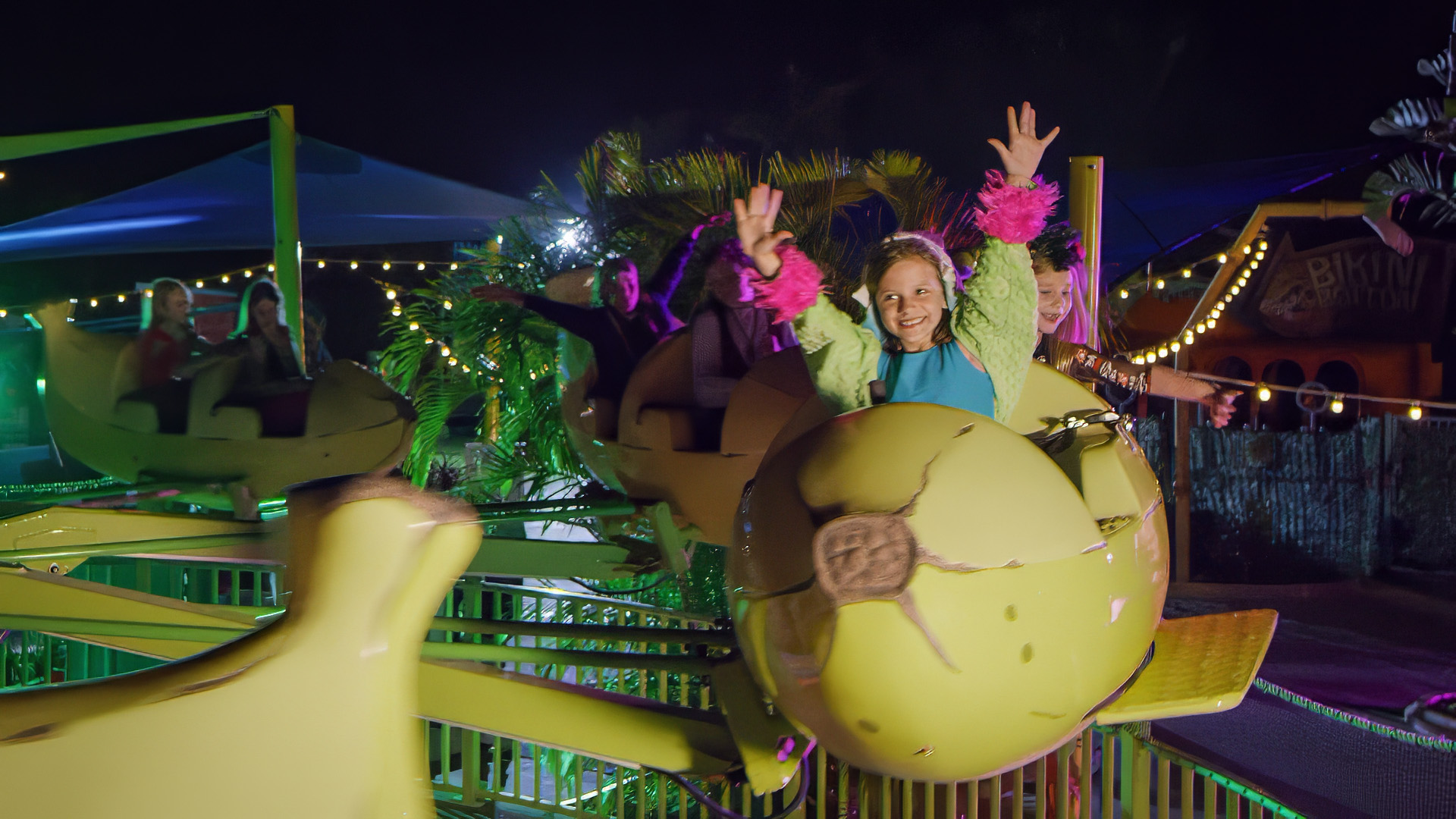 Children ride a yellow amusement park ride shaped like animals at night, smiling and raising their hands. The scene is lit with string lights and surrounded by greenery.