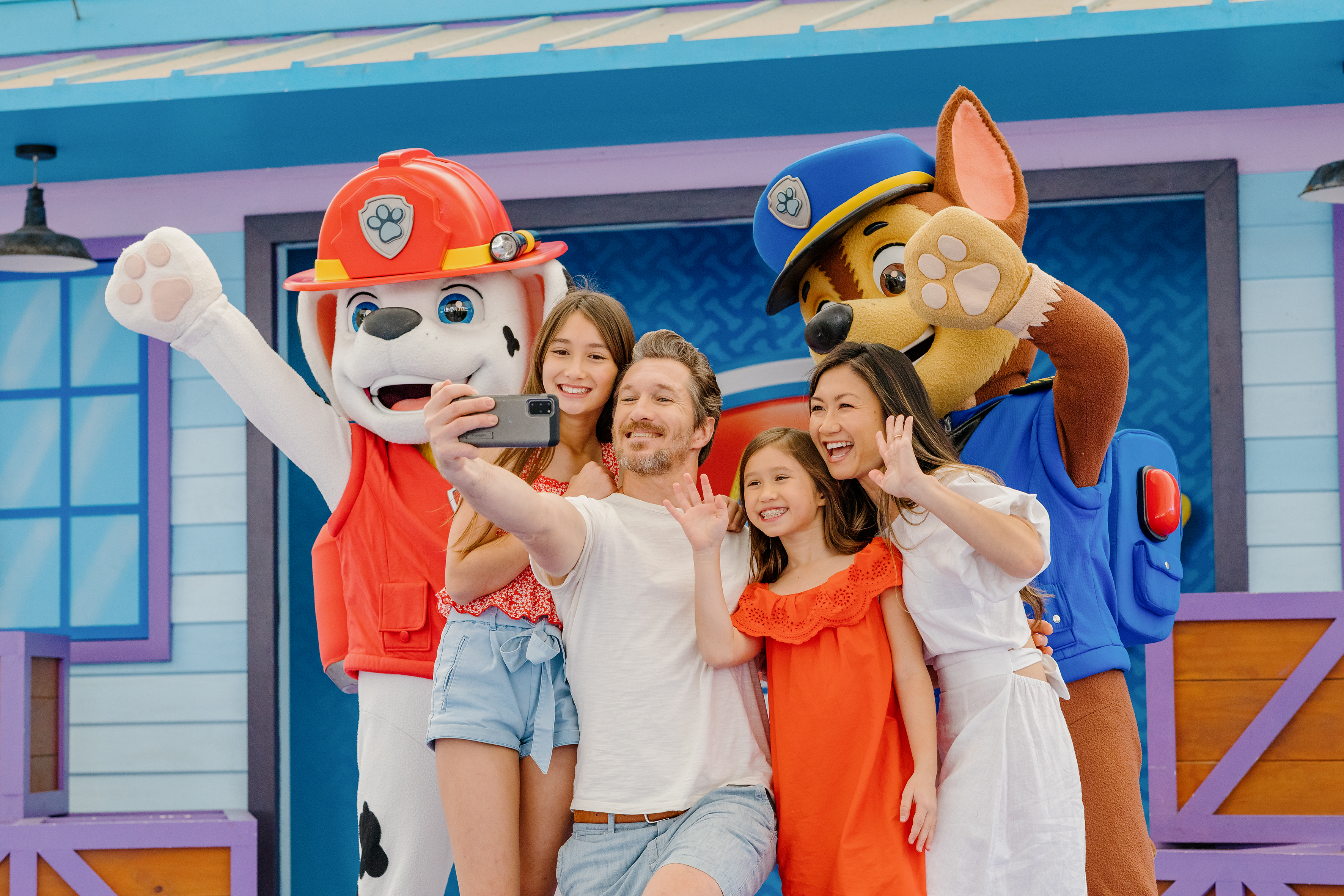 A family takes a selfie with two people dressed in Paw Patrol character costumes in front of a colorful building.