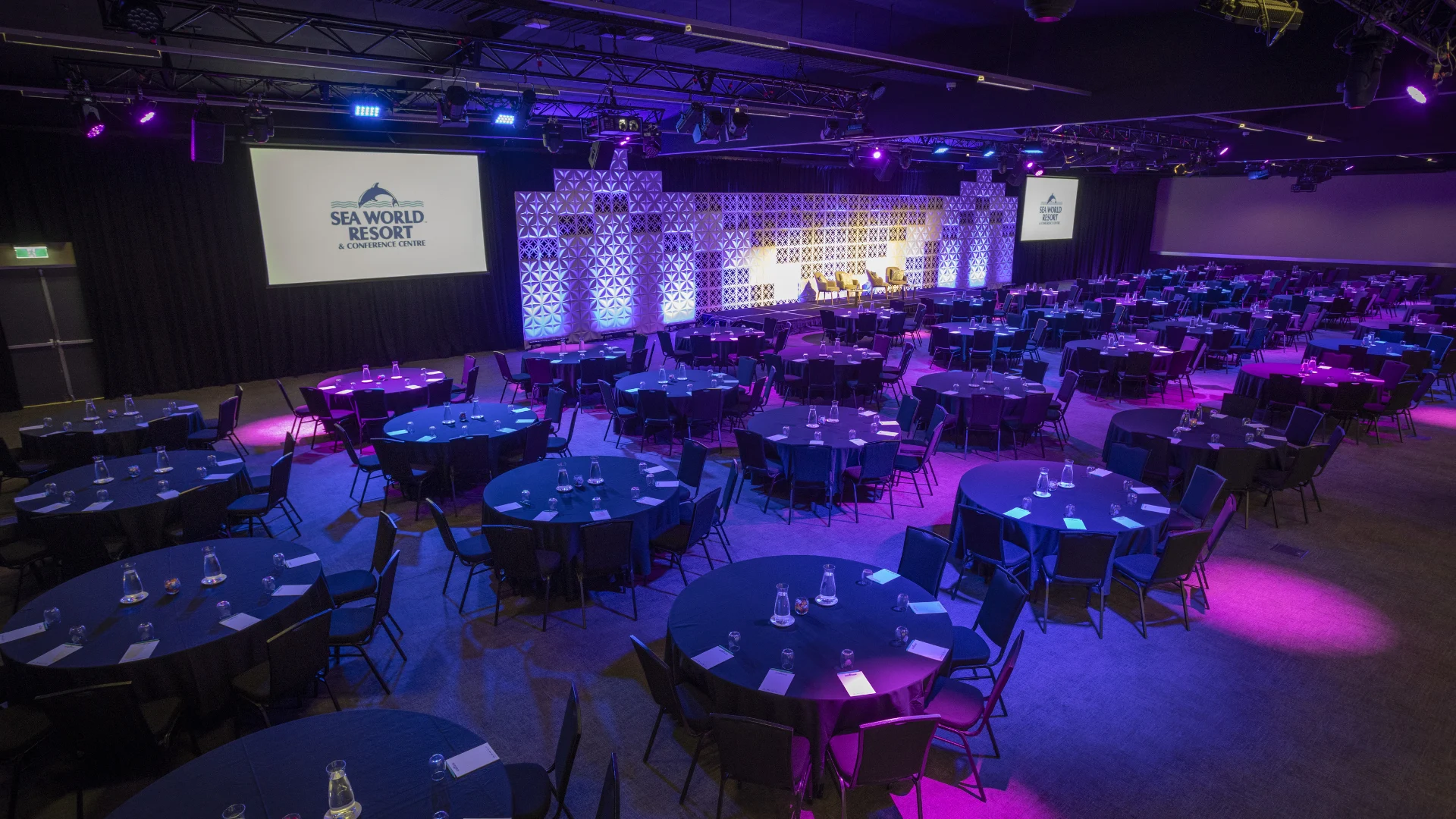 Large conference room with round tables and chairs set up, facing a stage with screens showing "Sea World Resort". Purple lighting illuminates the space.