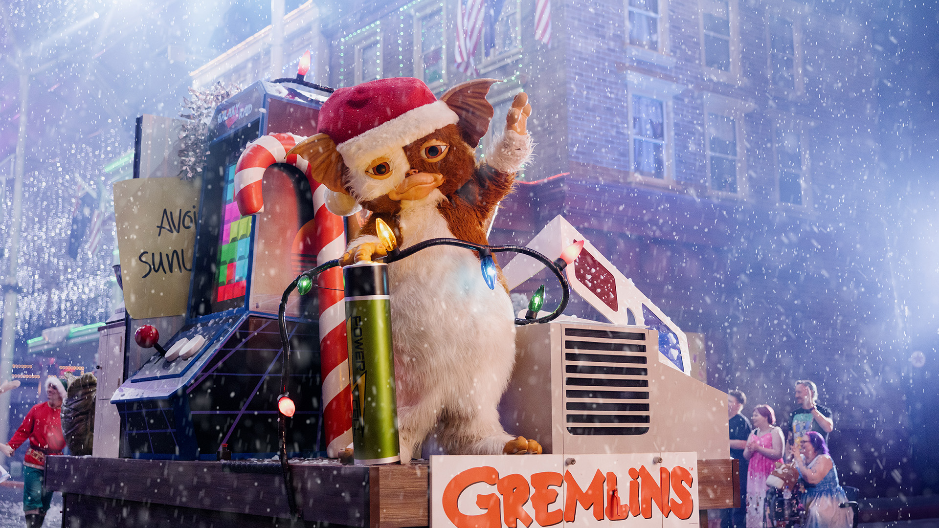 A furry creature wearing a Santa hat stands on a festive float with Christmas decorations, candy canes, and a sign that reads "Gremlins." Snow is falling, and people are visible in the background on a city street.