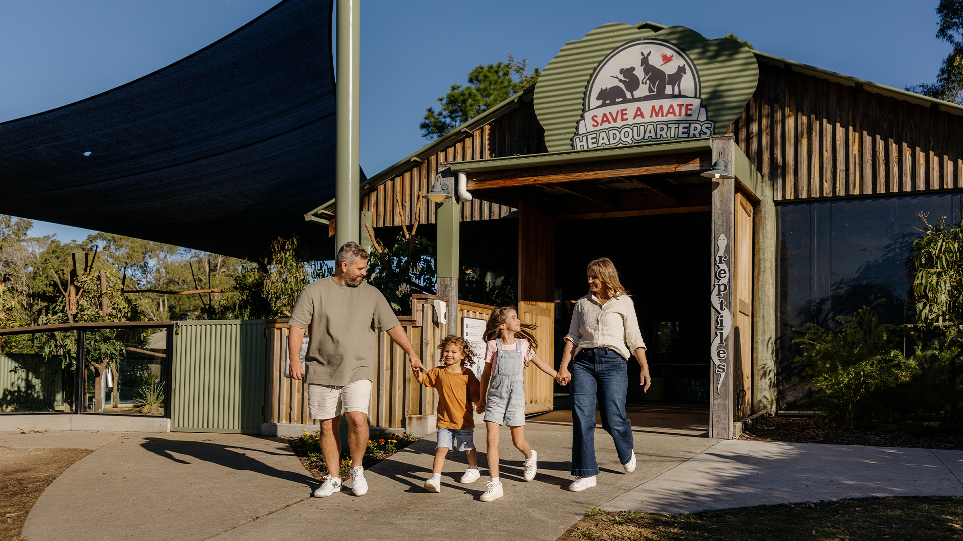A family of four, two adults and two children, walk hand in hand outside a rustic building labeled "Save a Mate Headquarters" on a sunny day, surrounded by greenery.