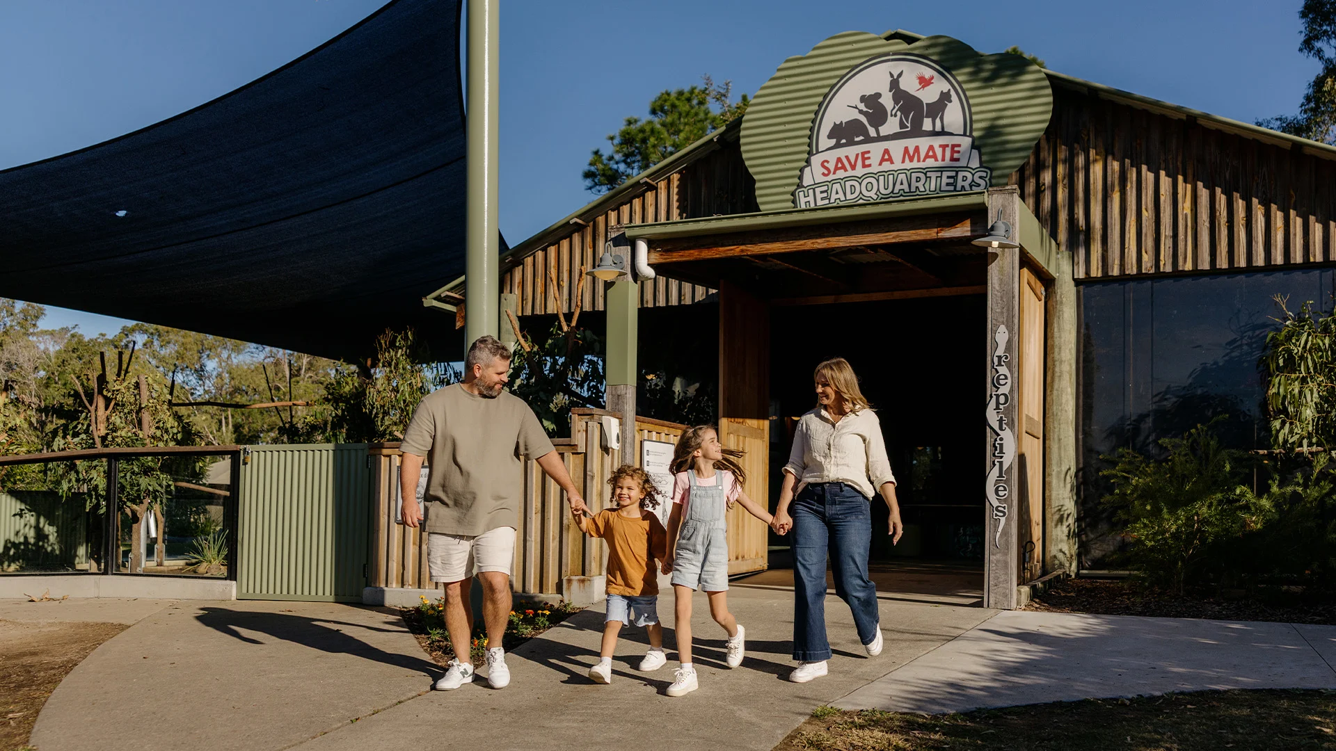 A family of four, two adults and two children, walk hand in hand outside a rustic building labeled "Save a Mate Headquarters" on a sunny day, surrounded by greenery.