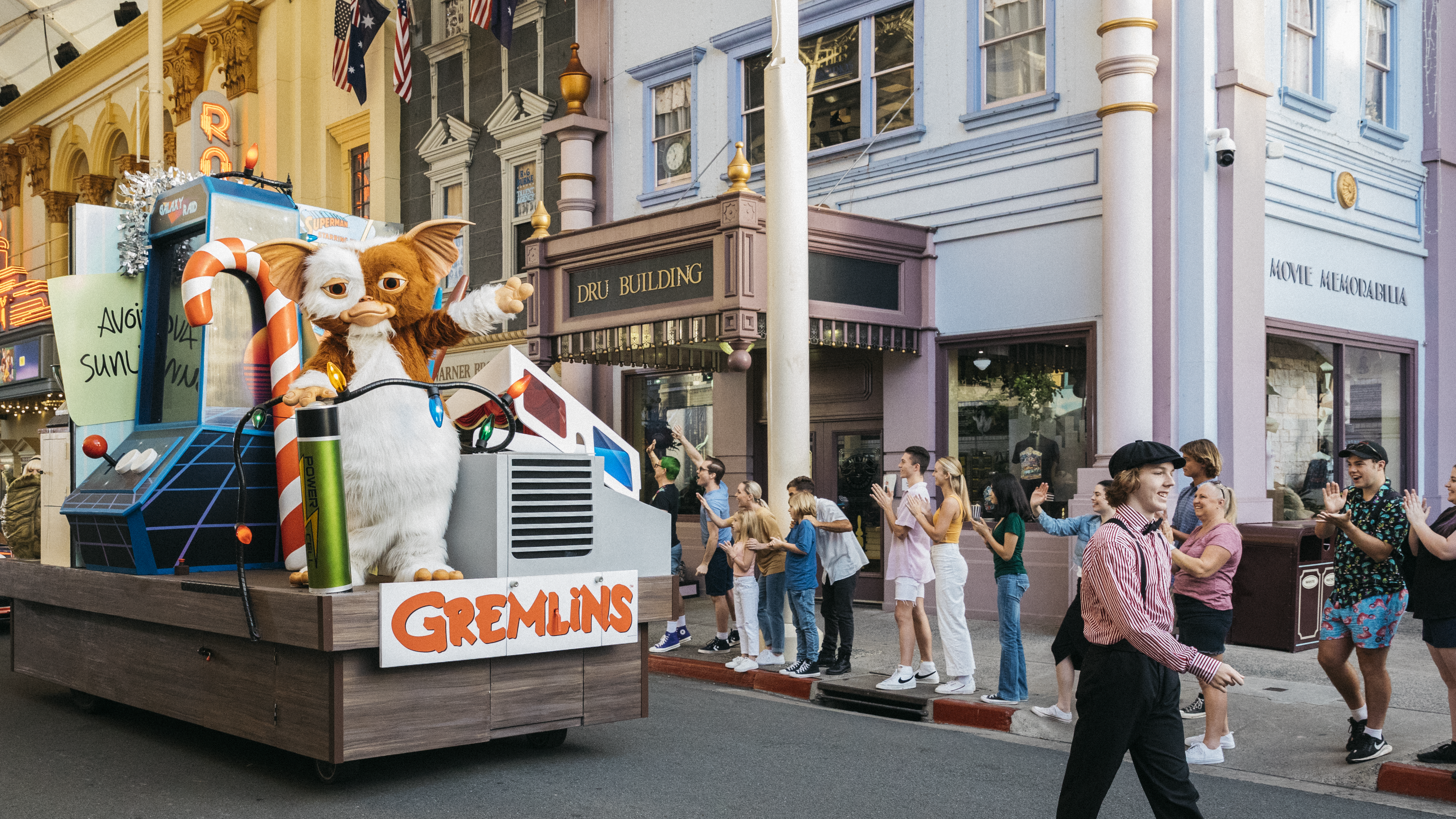 A parade float themed after "Gremlins," featuring large Gizmo and Stripe figures, moves down a street as spectators on the sidewalk watch and take photos in a lively, amusement park setting.