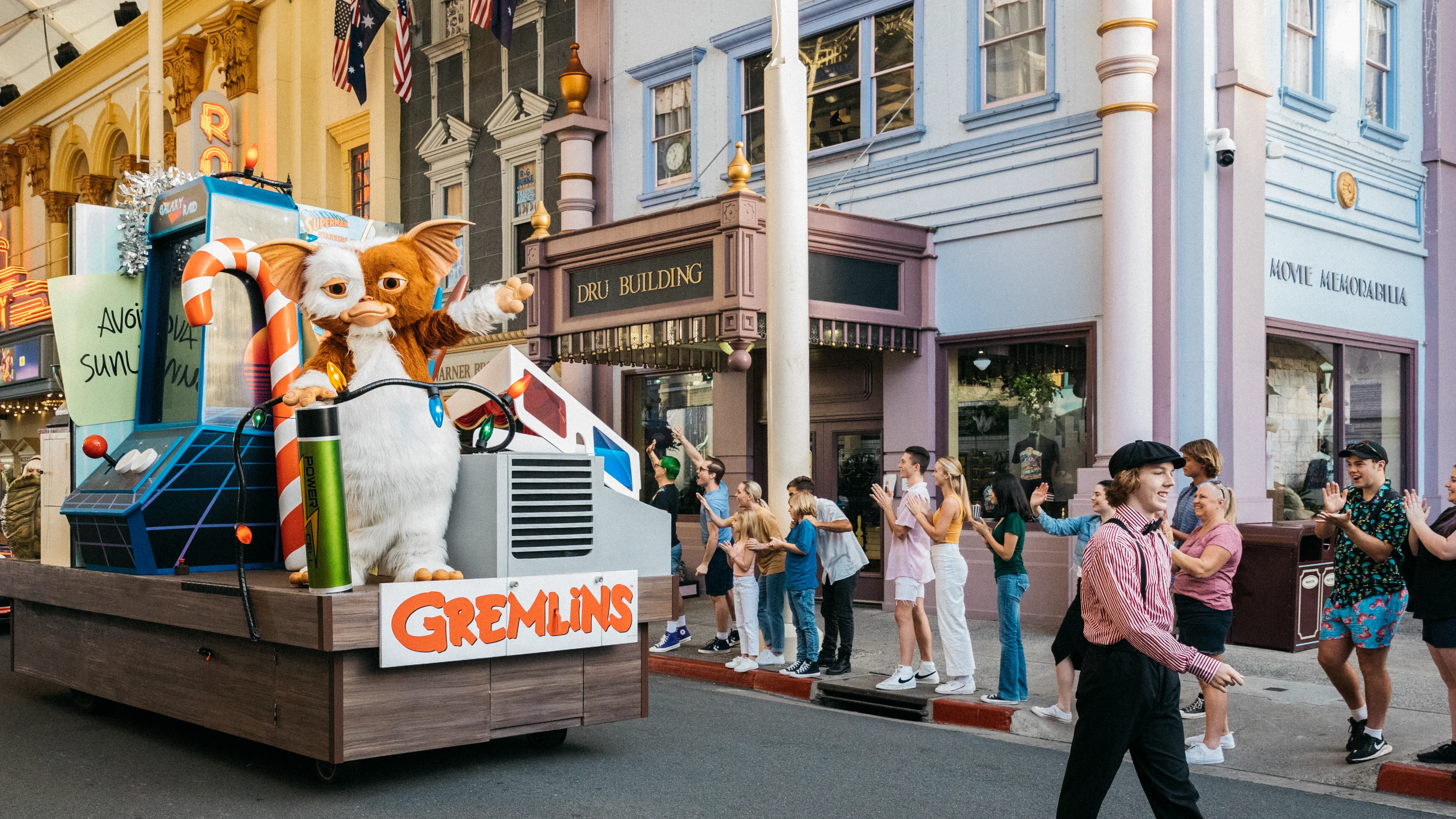 A parade float themed after "Gremlins," featuring large Gizmo and Stripe figures, moves down a street as spectators on the sidewalk watch and take photos in a lively, amusement park setting.
