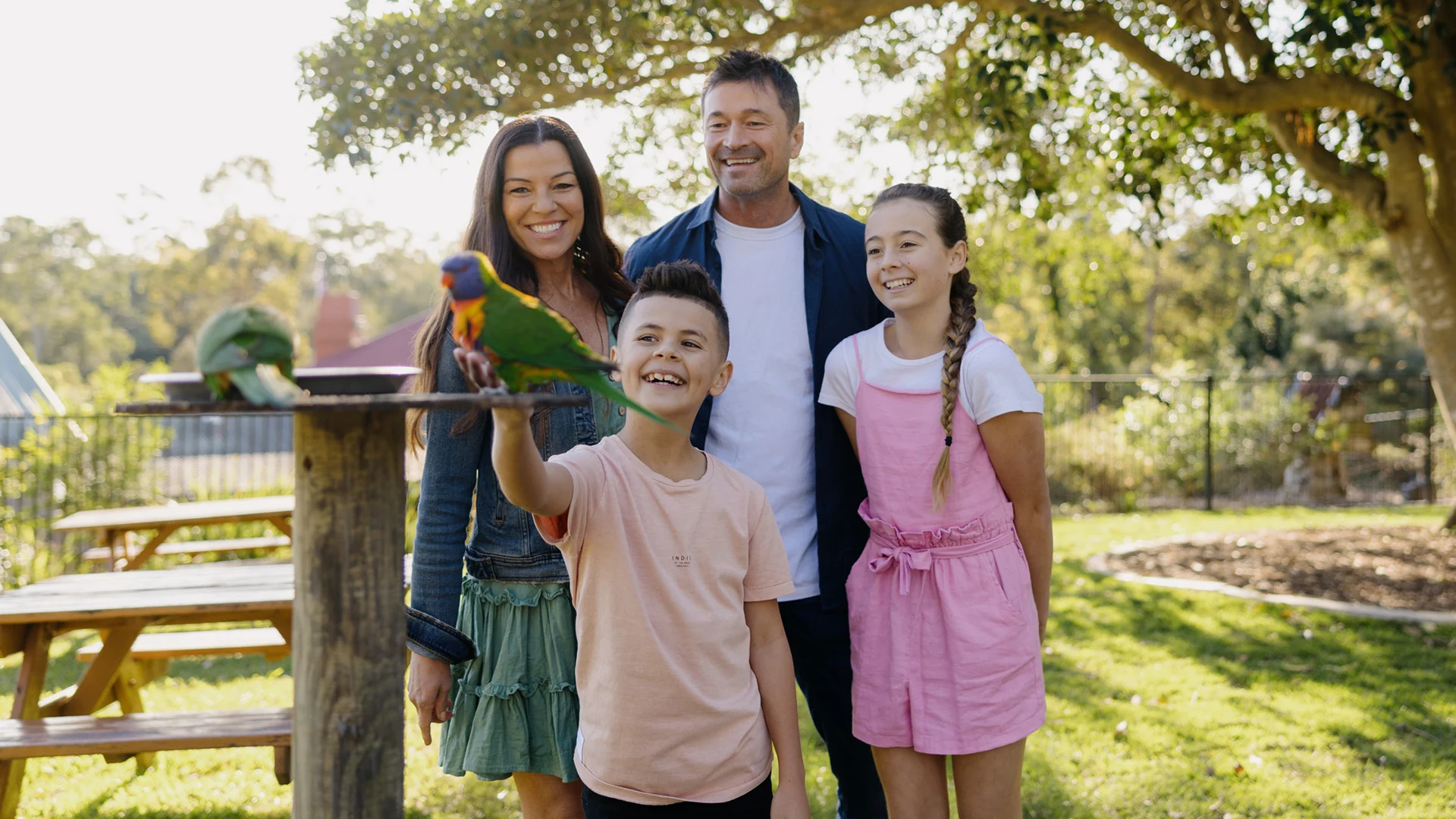 Two young girls hold a metal dish and smile while a colorful parrot perches on the edge, with greenery in the background.
