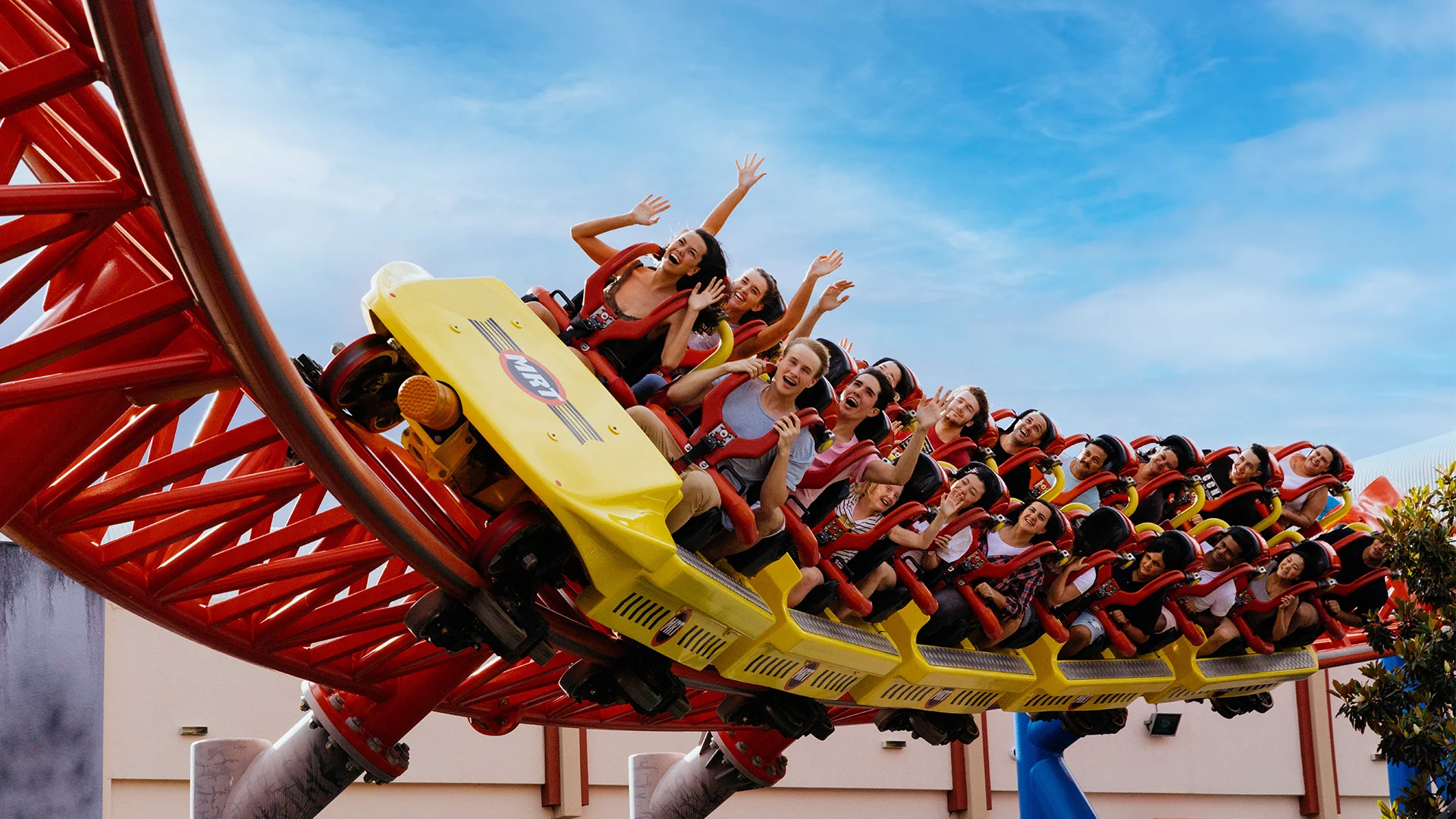 A group of people riding a red and yellow roller coaster, some with their hands raised and expressions of excitement as it rounds a curve under a blue sky.