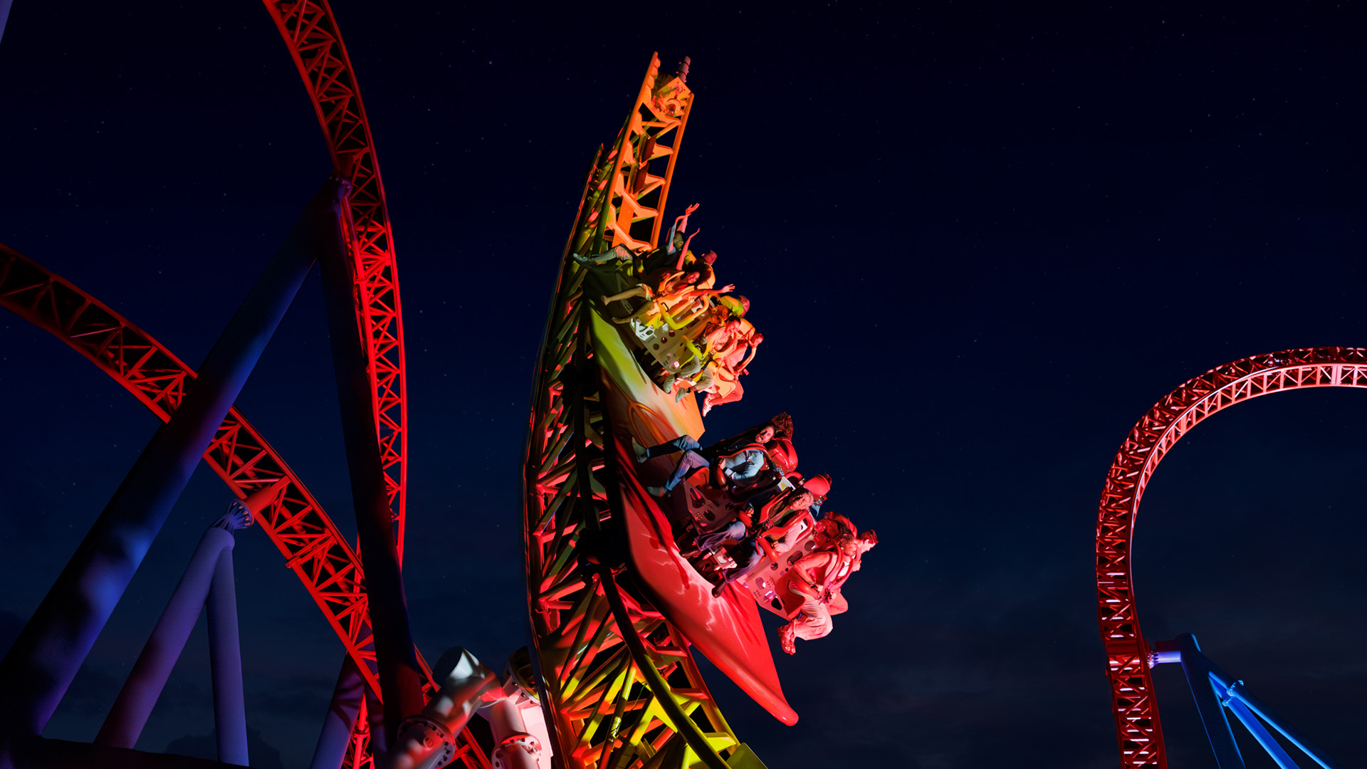 A brightly lit roller coaster with riders speeding downward at night, arms raised and faces excited, surrounded by red and yellow tracks against a dark sky.