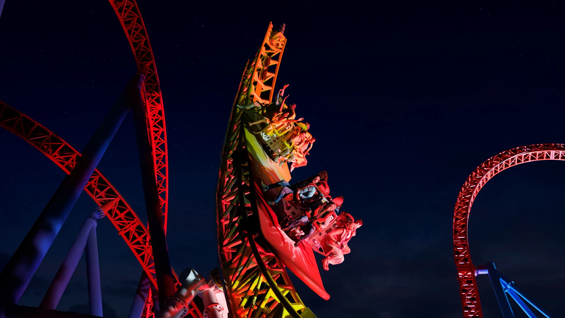 A brightly lit roller coaster with riders speeding downward at night, arms raised and faces excited, surrounded by red and yellow tracks against a dark sky.