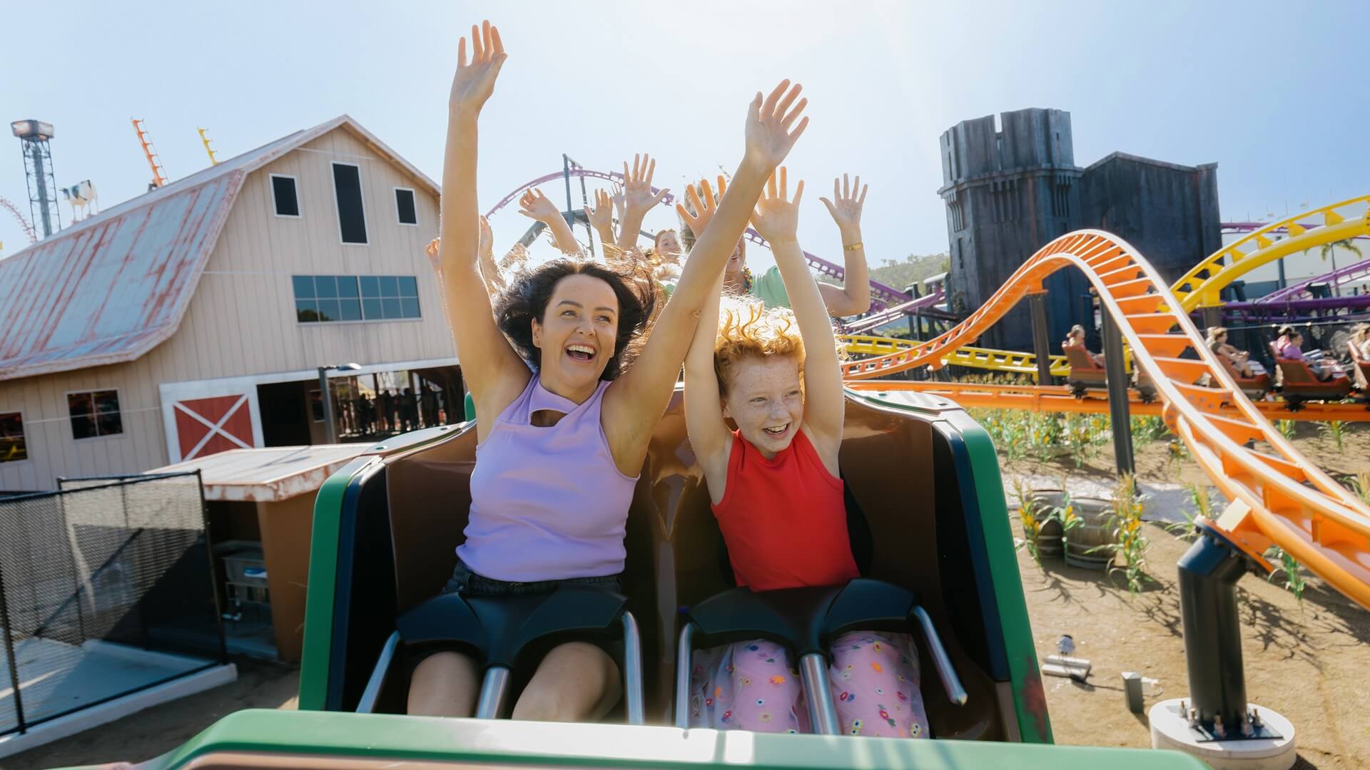 Excited mother and daughter riding a colourful outdoor roller coaster with arms raised, laughing as the ride twists through bright orange tracks at a theme park