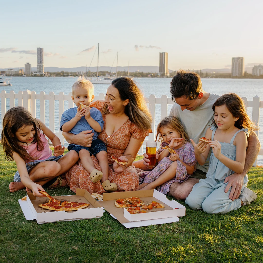 A family of six sits on grass by the water, eating pizza from open boxes. The sun is setting, and city buildings and boats are visible in the background. Everyone looks happy and relaxed, enjoying their picnic.
