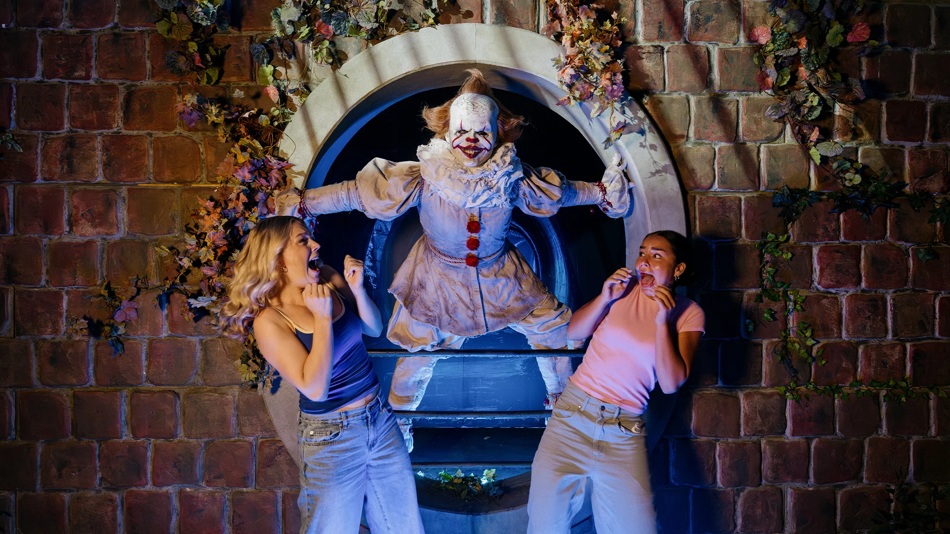 Two frightened women recoil as a clown in a ruffled costume and white makeup lunges from a circular stone tunnel surrounded by vines.