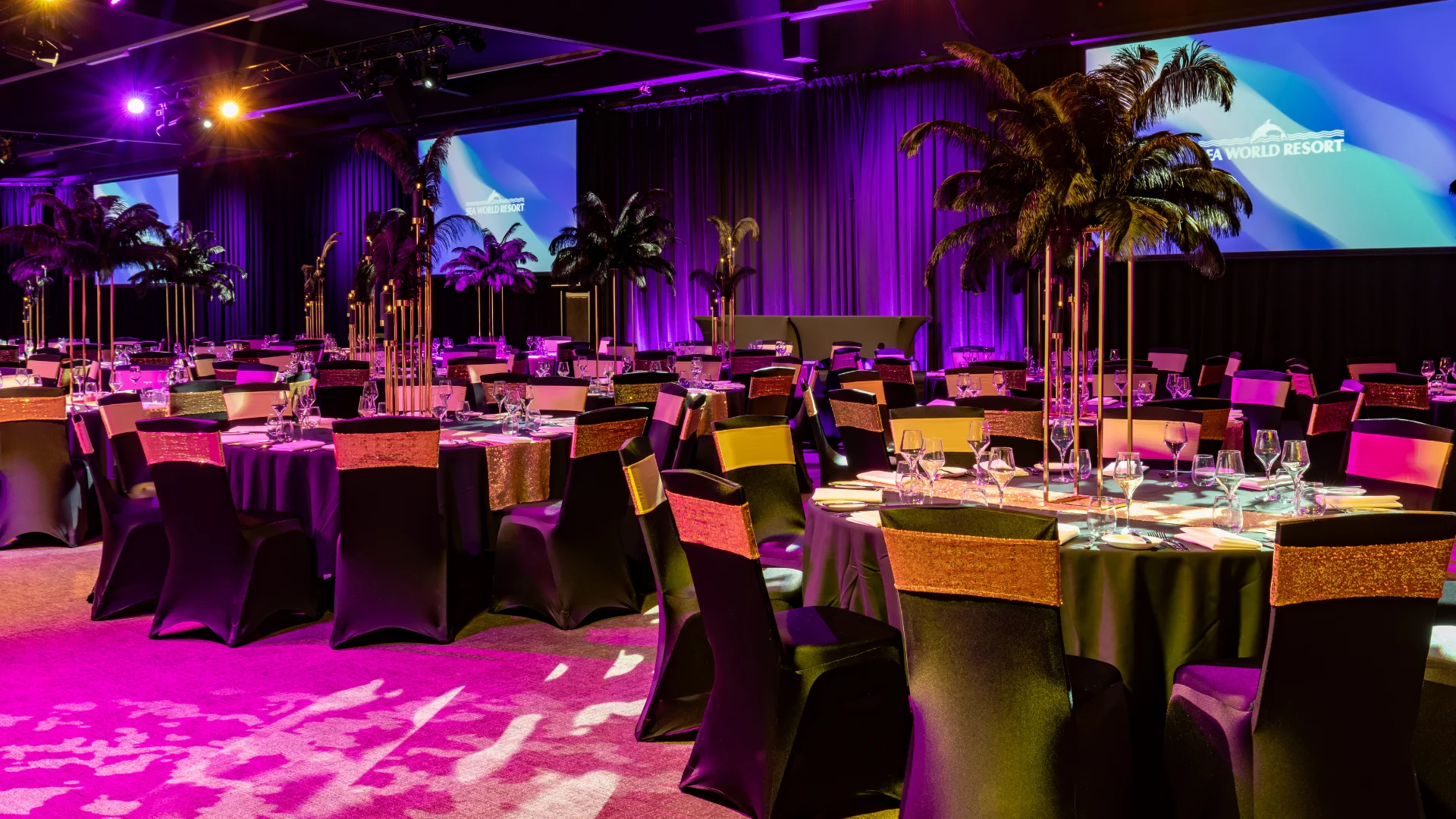 A banquet hall with round tables covered in black linens, tall palm centerpieces, and elegant place settings, lit by purple and gold lights. Two large screens display colorful graphics at the front of the room.