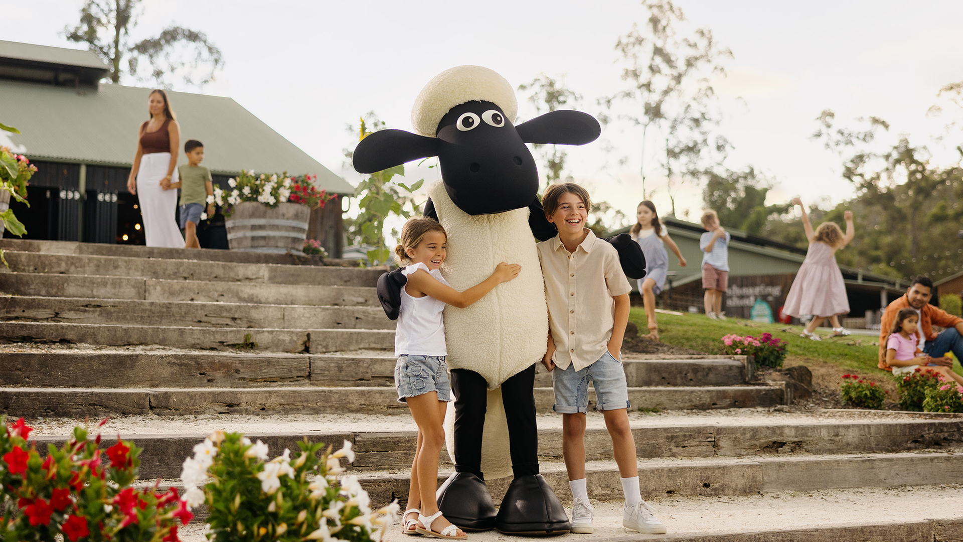Two children hug a person in a large Shaun the Sheep costume on stone steps outdoors, with colorful flowers nearby and several people in the background enjoying a sunny day.