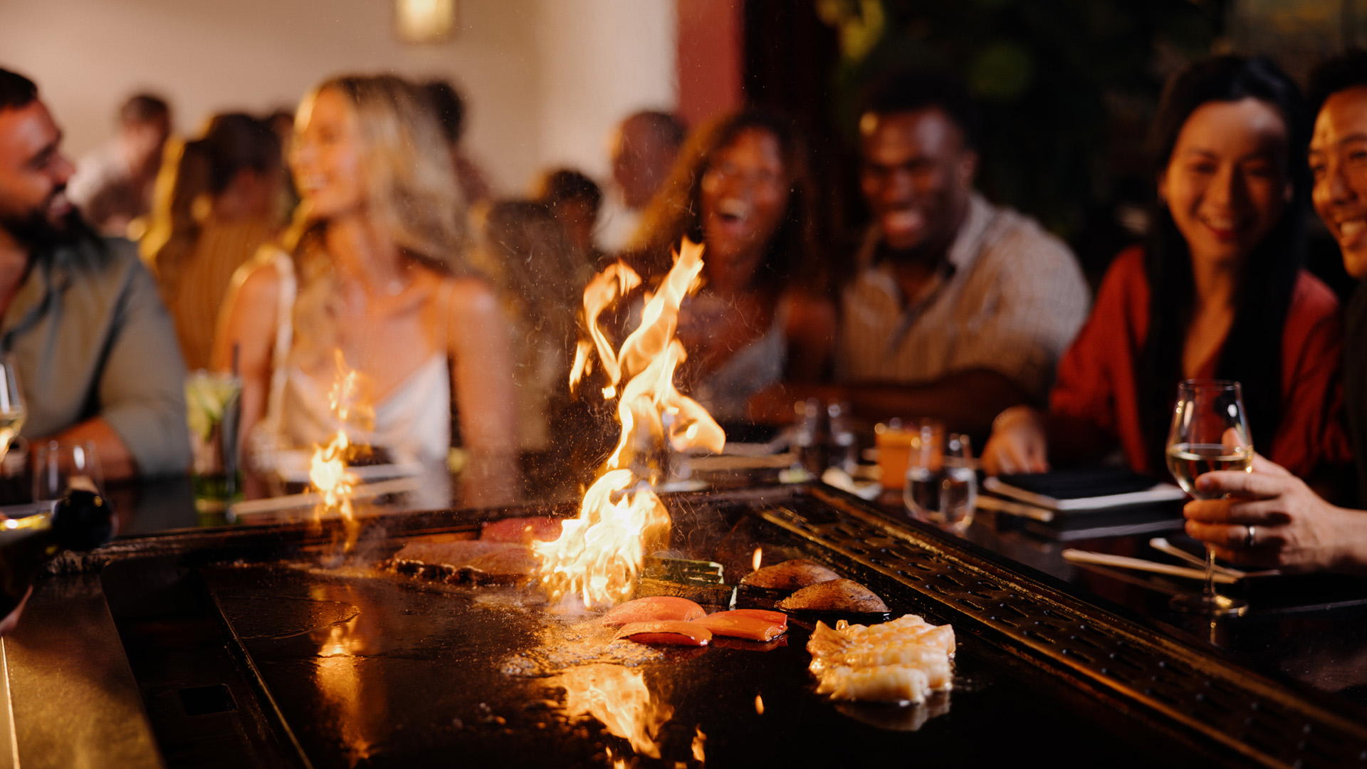 A group of people sit around a teppanyaki grill at a restaurant, smiling and enjoying themselves as a chef cooks food with flames rising from the hot grill in the foreground.