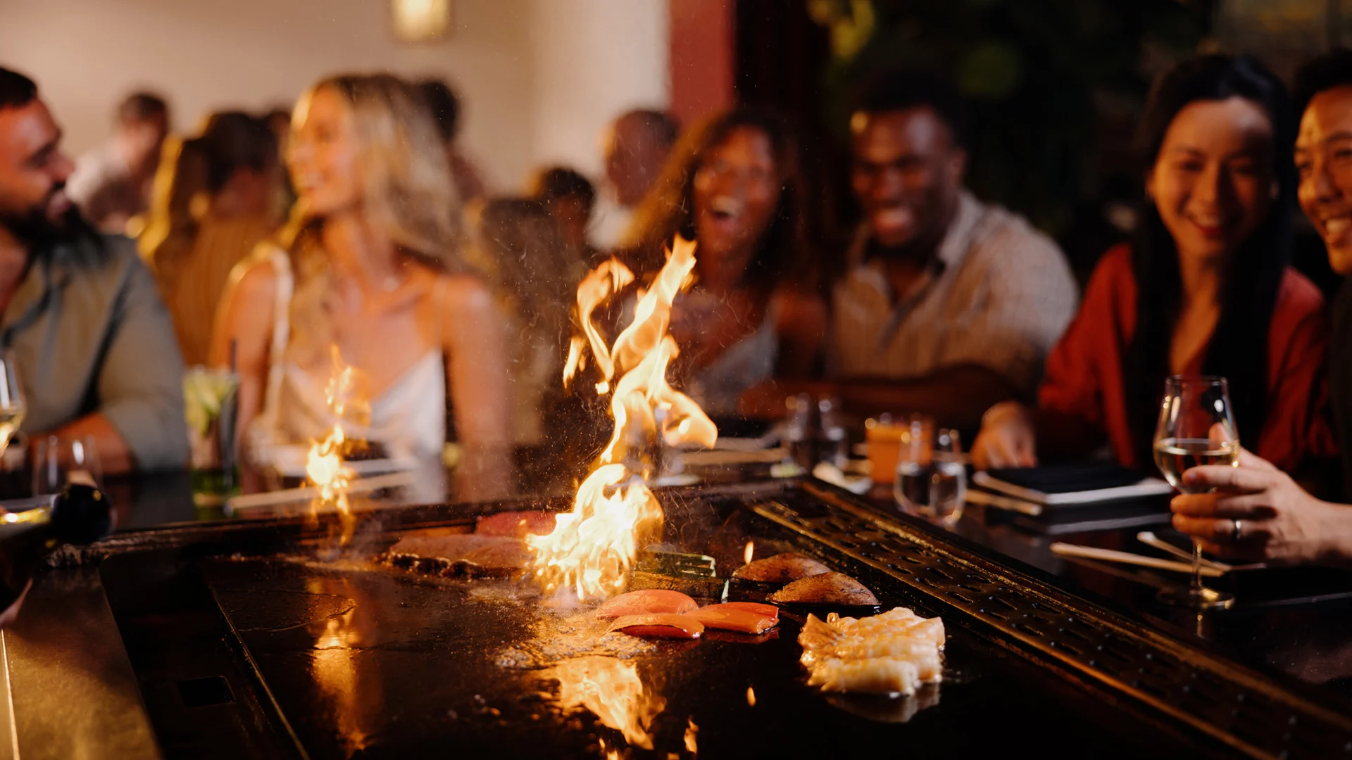 A group of people sit around a teppanyaki grill at a restaurant, smiling and enjoying themselves as a chef cooks food with flames rising from the hot grill in the foreground.