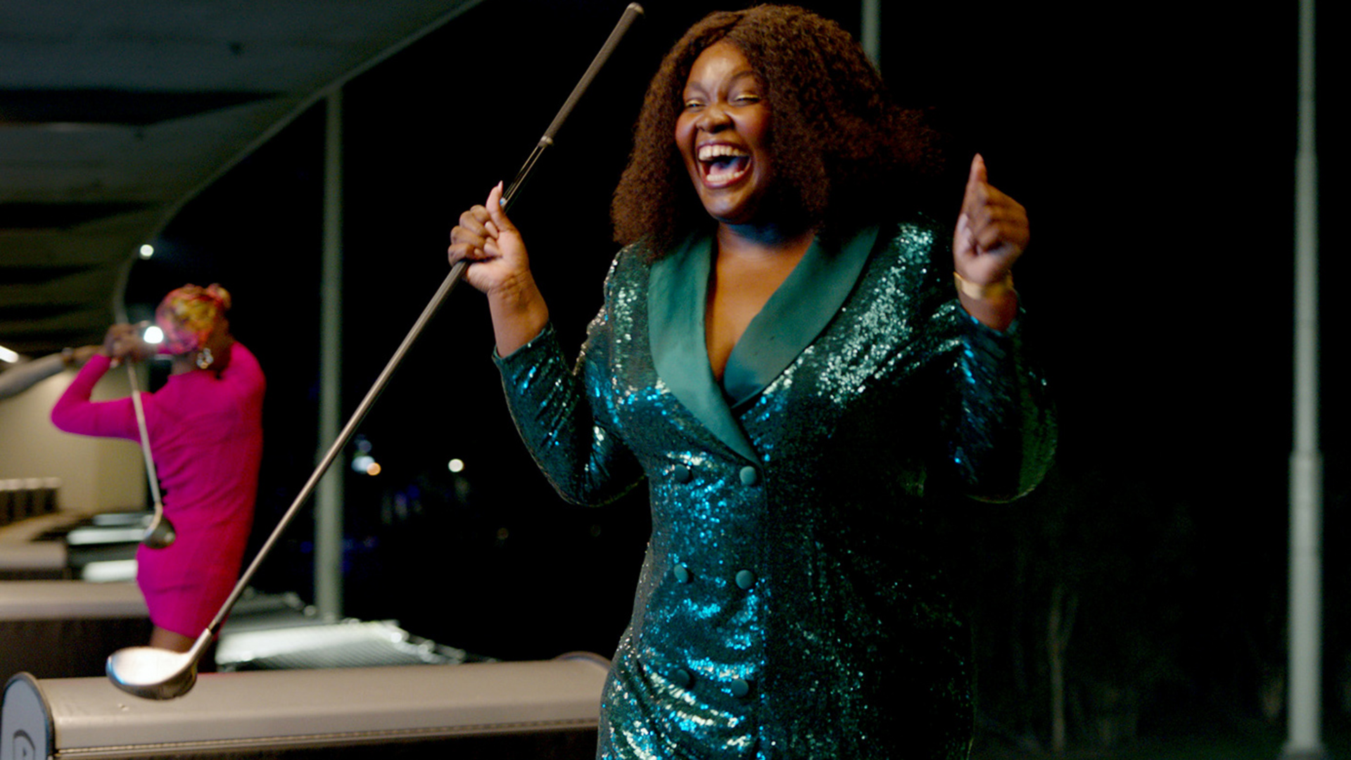 A woman in a sparkly green dress smiles and cheers while holding a golf club. Another person in pink swings a club in the background. They appear to be at a driving range at night.