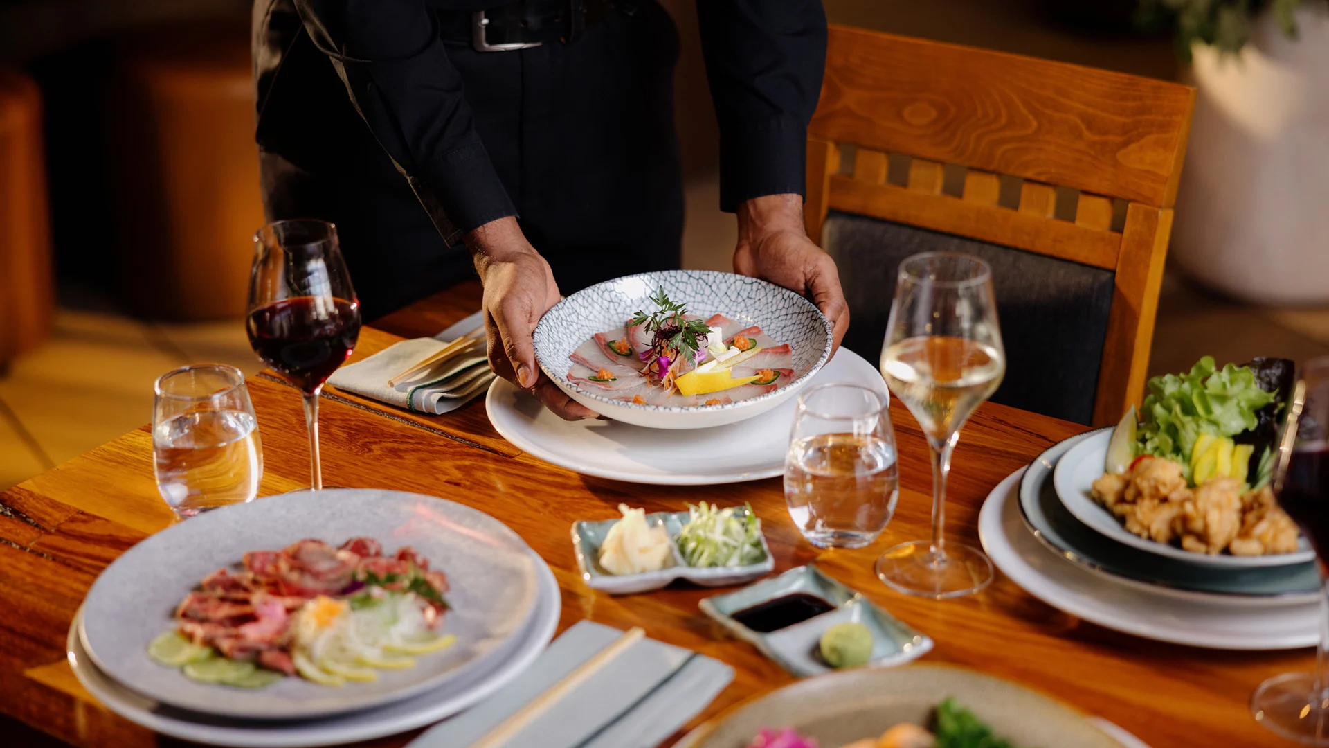 A server places a plated dish on a wooden table, surrounded by wine glasses and various dishes, including salad and sashimi, at Hatsuhana Japanese Restaurant.