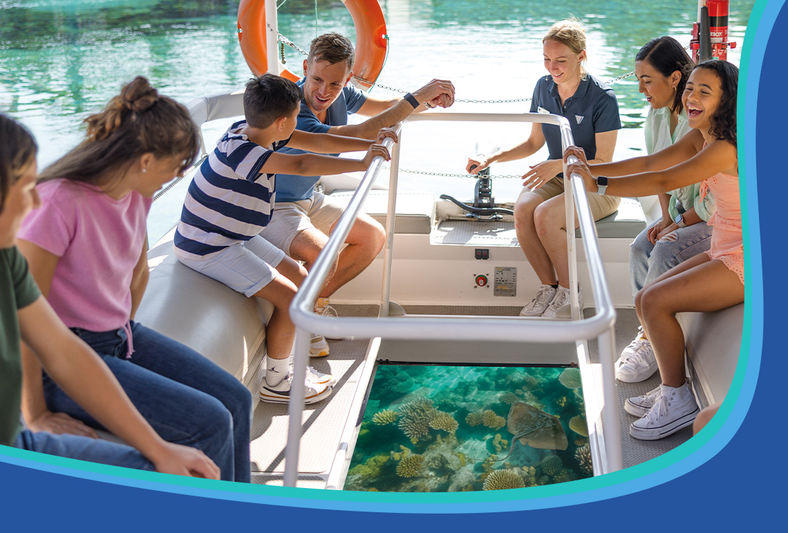 A group of people sit on a glass-bottom boat, looking down at coral reefs in the clear water below while a guide speaks to them.