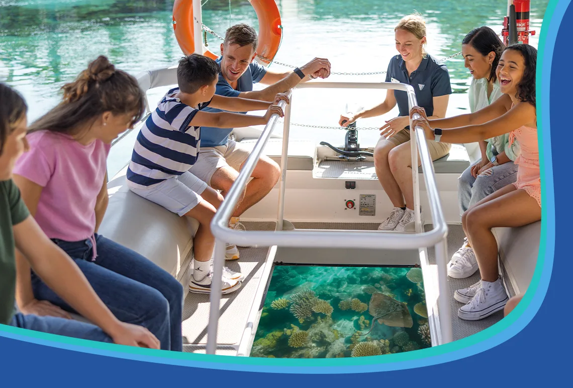 A group of people sit on a glass-bottom boat, looking down at coral reefs in the clear water below while a guide speaks to them.