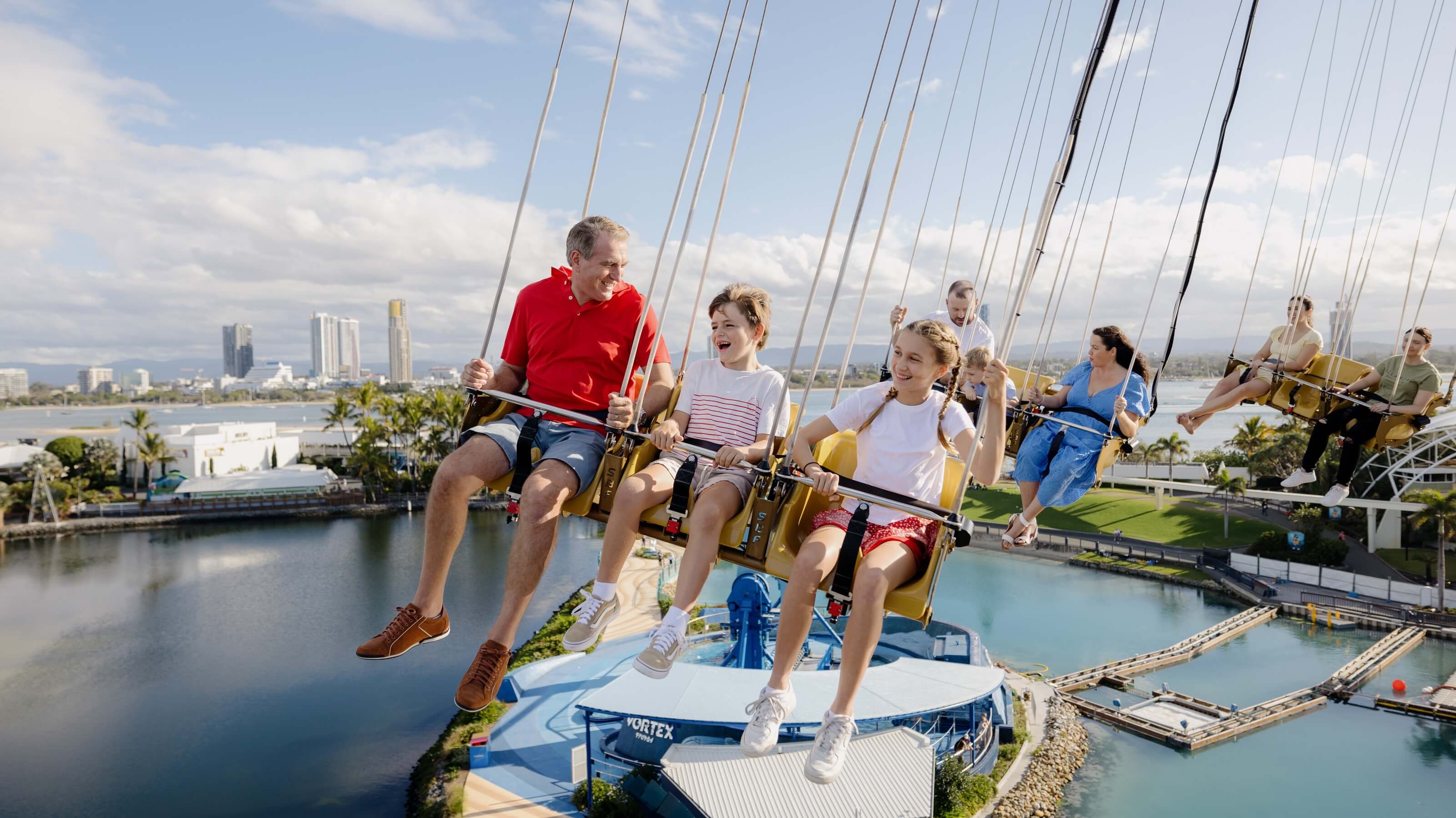Visitors revel in the thrill of a swing ride, Trident, soaring above glistening waters at Sea World on the Gold Coast. The charming backdrop of buildings and a cloudy sky adds to the enchanting experience.