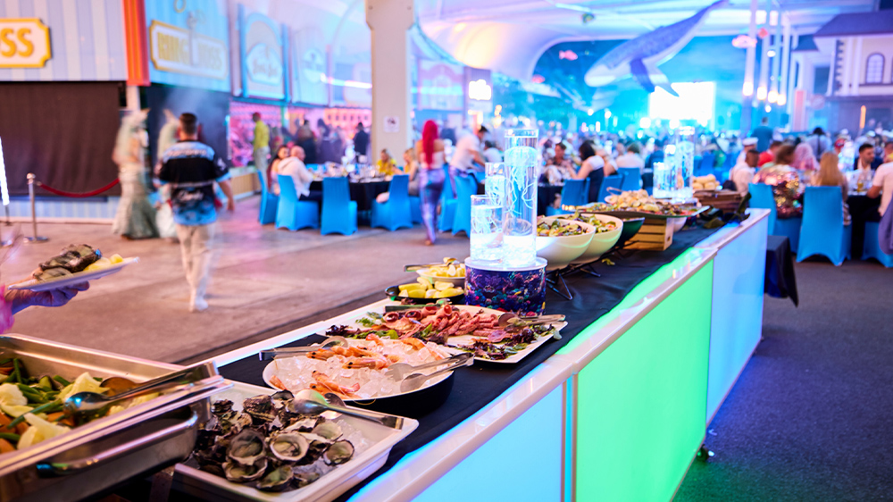A buffet table with assorted food is set up at a lively, brightly lit indoor event with many people seated at tables; a large shark decoration hangs from the ceiling in the background.