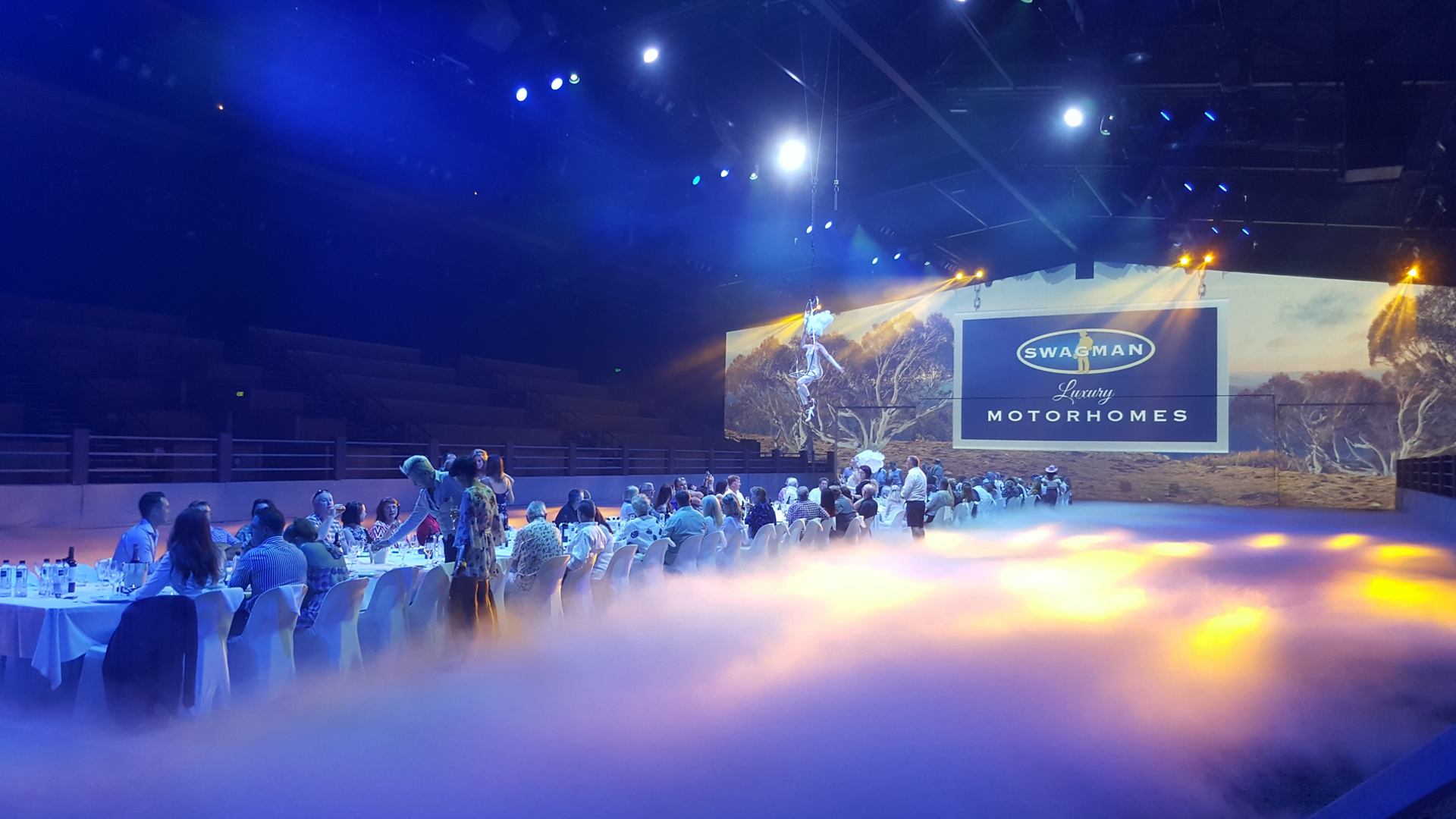 A large group of people sit at a long banquet table in a dimly lit venue with dramatic stage lighting and fog effects. A screen in the background displays "SWANMAN Luxury MOTORHOMES.