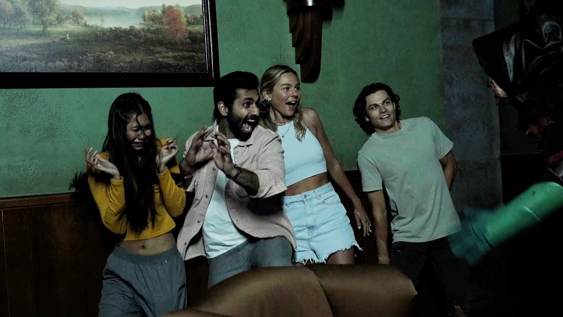 Four young adults stand close together indoors, reacting with surprise and fear to something off-camera. Their expressions and body language suggest they are startled or scared.