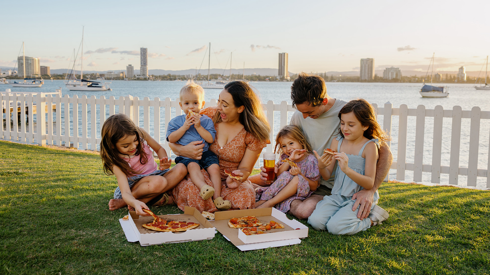 A family of six sits on grass by a waterfront, enjoying pizza together at sunset. There are boats in the water and tall buildings in the background, with a white picket fence behind them.