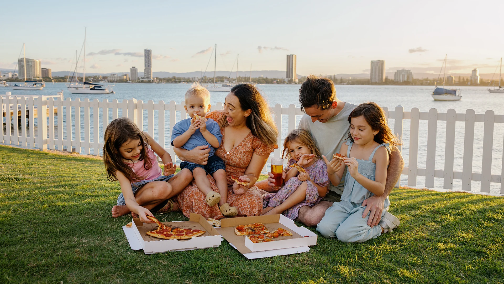 A family of six sits on grass by a waterfront, enjoying pizza together at sunset. There are boats in the water and tall buildings in the background, with a white picket fence behind them.
