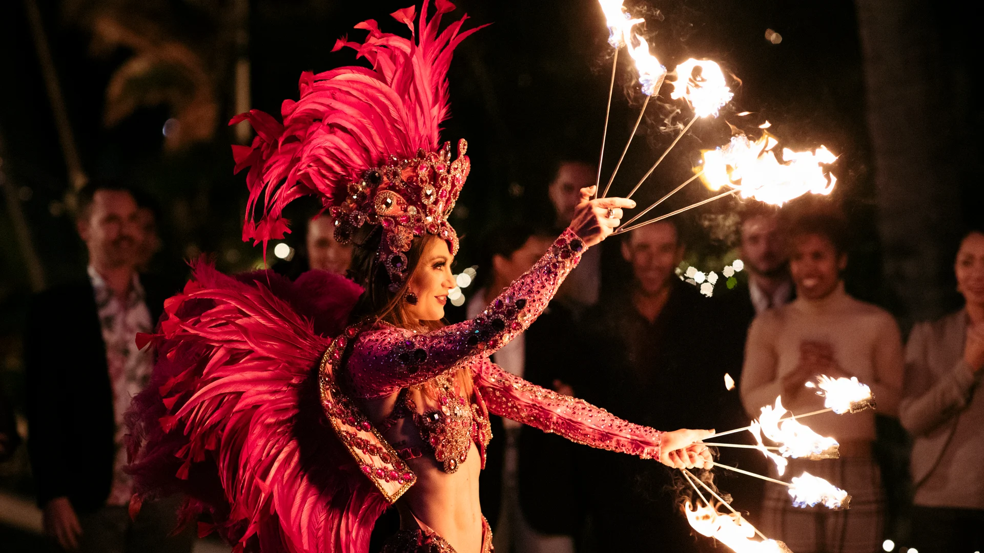 A performer in a vibrant red feathered costume and headdress entertains a crowd at night, skillfully spinning fire sticks, while onlookers watch and smile in the background.