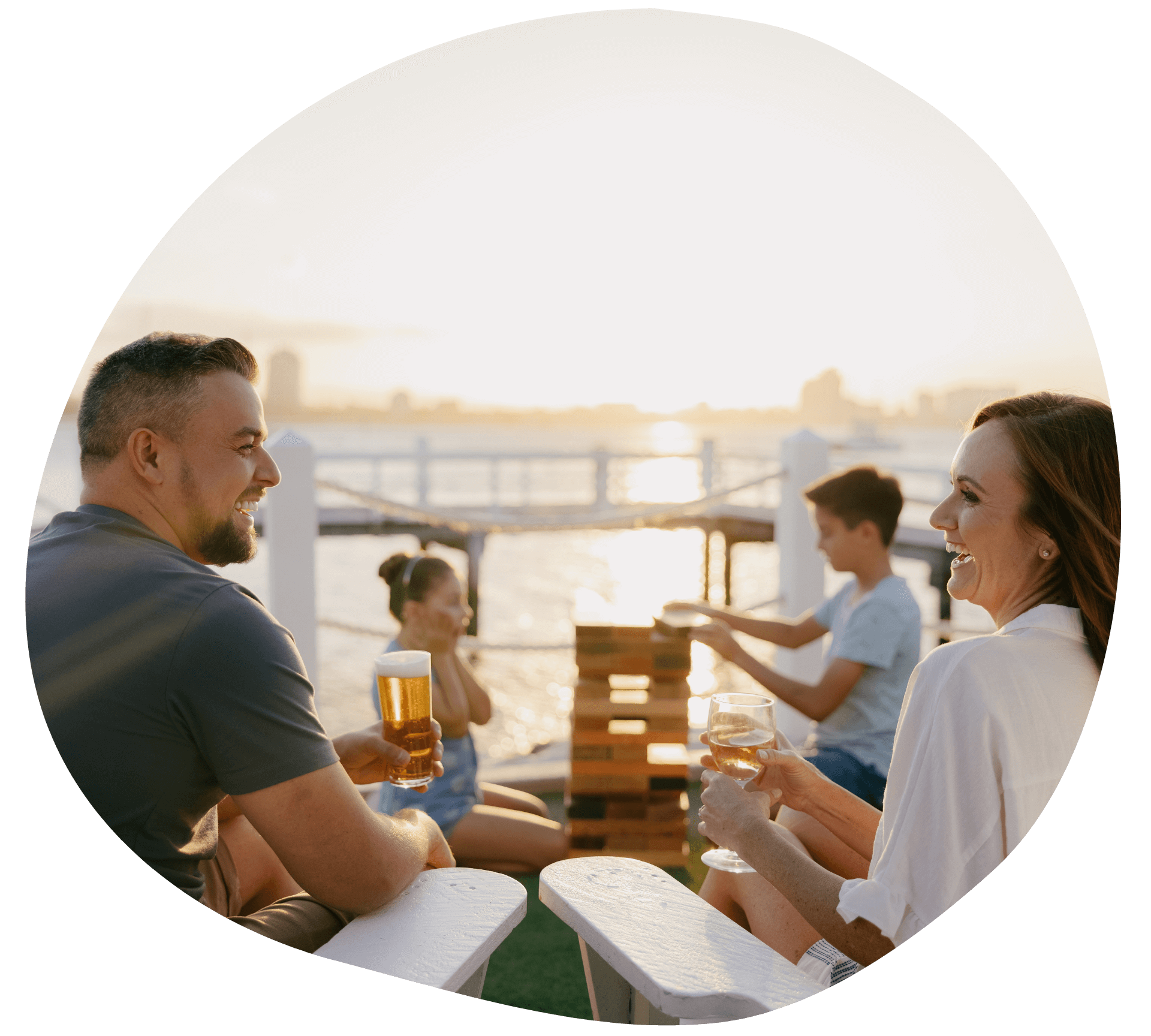 Four people sitting outdoors on a deck by the water, playing Jenga and enjoying drinks, with a sunset in the background. Located at the Boatshed, Sea World Resort.