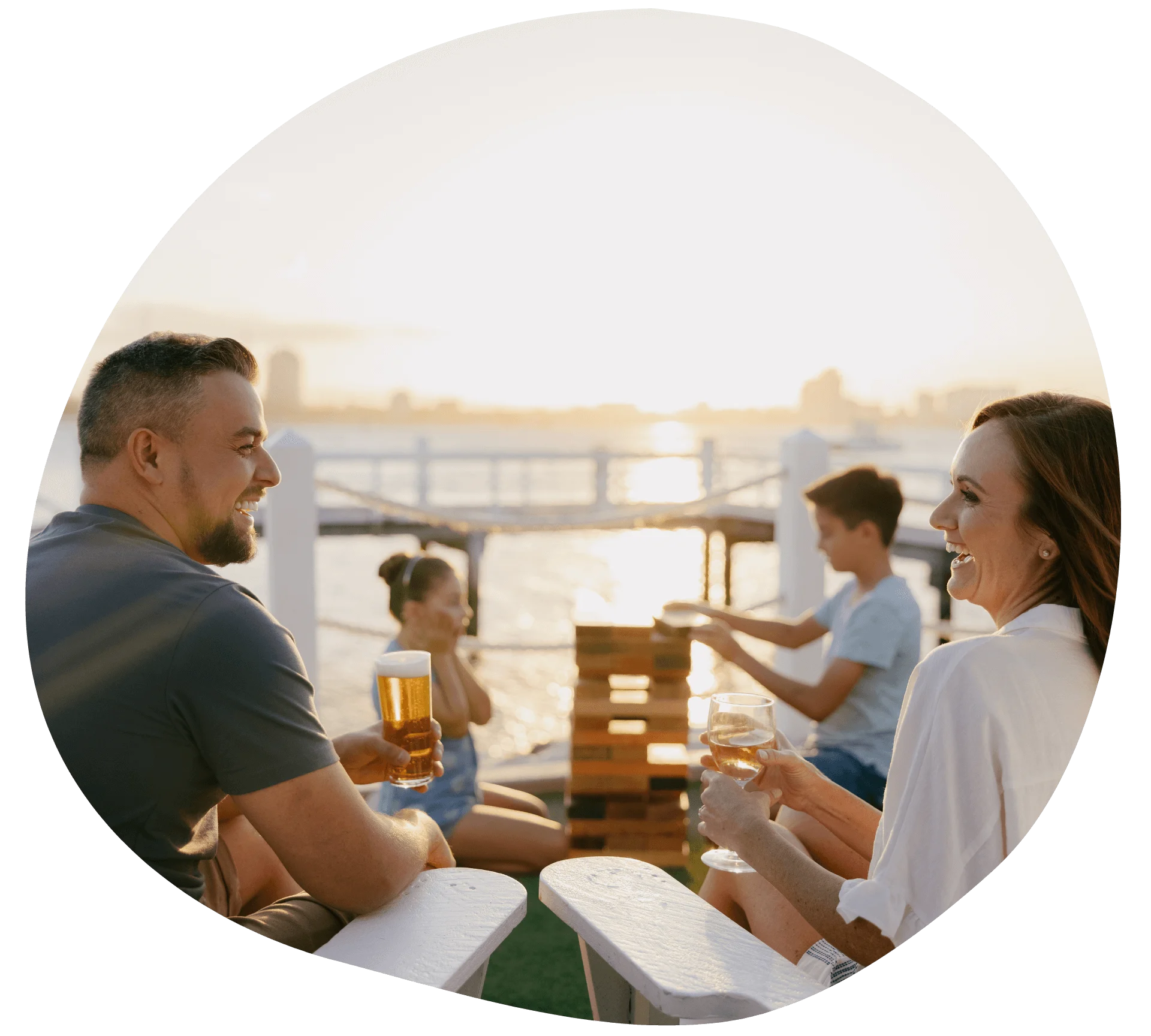 Four people sitting outdoors on a deck by the water, playing Jenga and enjoying drinks, with a sunset in the background. Located at the Boatshed, Sea World Resort.