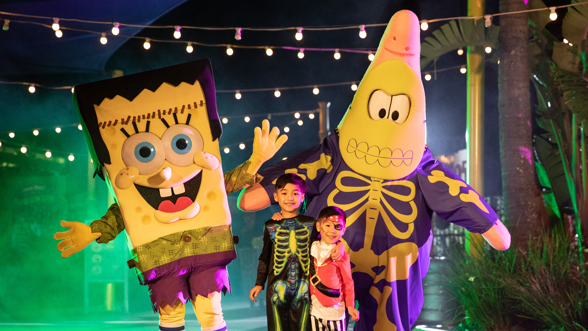 Two children in Halloween costumes stand between people dressed as SpongeBob and Patrick in skeleton outfits, under string lights at night with colorful fog and palm trees in the background.