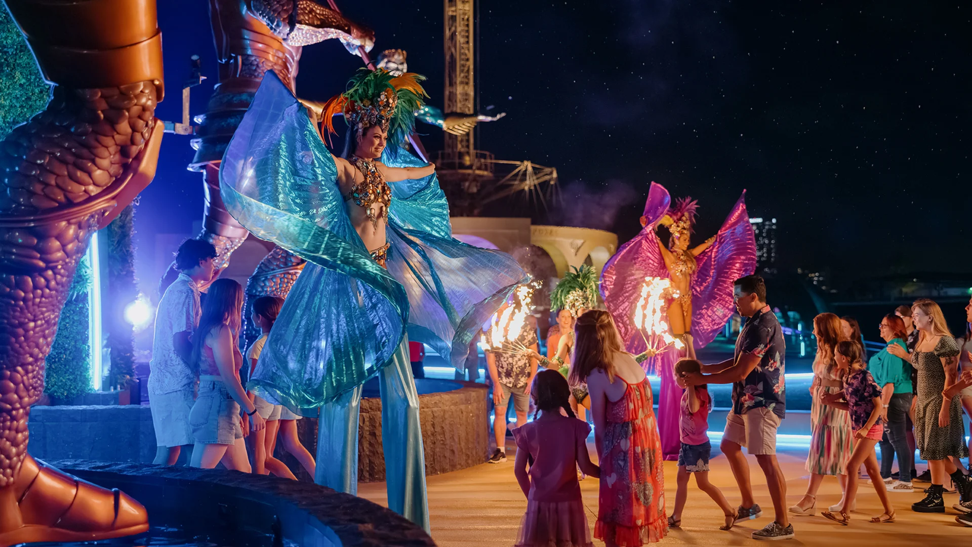 Performers in colorful, elaborate costumes on stilts entertain a group of families with children at night, under bright lights, in an outdoor festive setting.