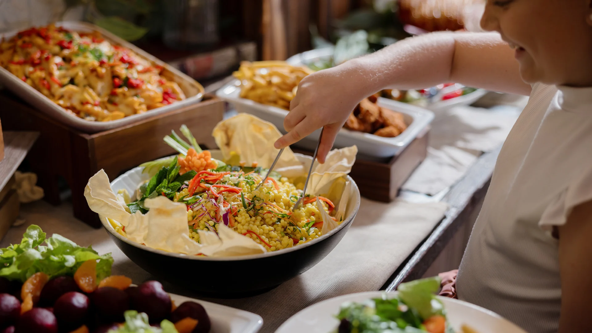 A person serving themselves salad from a large bowl at a buffet table with various dishes and trays of food.