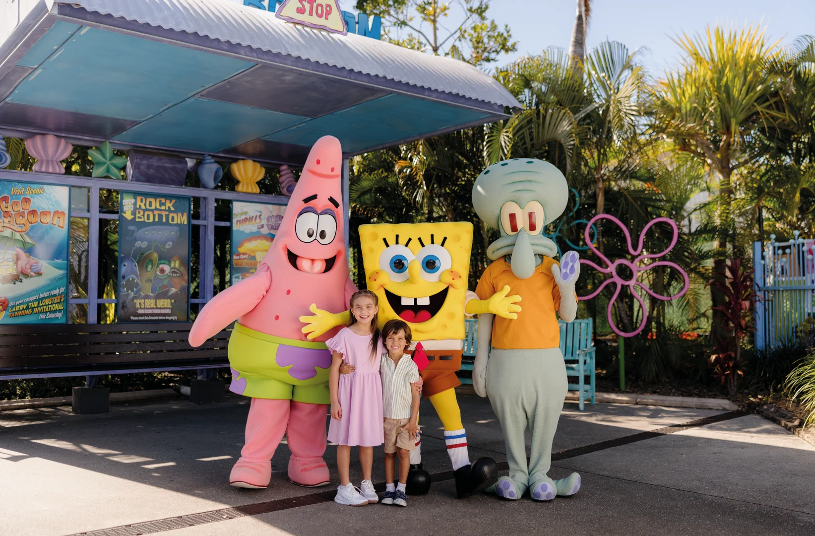 Two young children stand smiling with three people in costumes of Patrick Star, SpongeBob SquarePants, and Squidward under an outdoor pavilion, surrounded by palm trees and colorful theme park decorations.