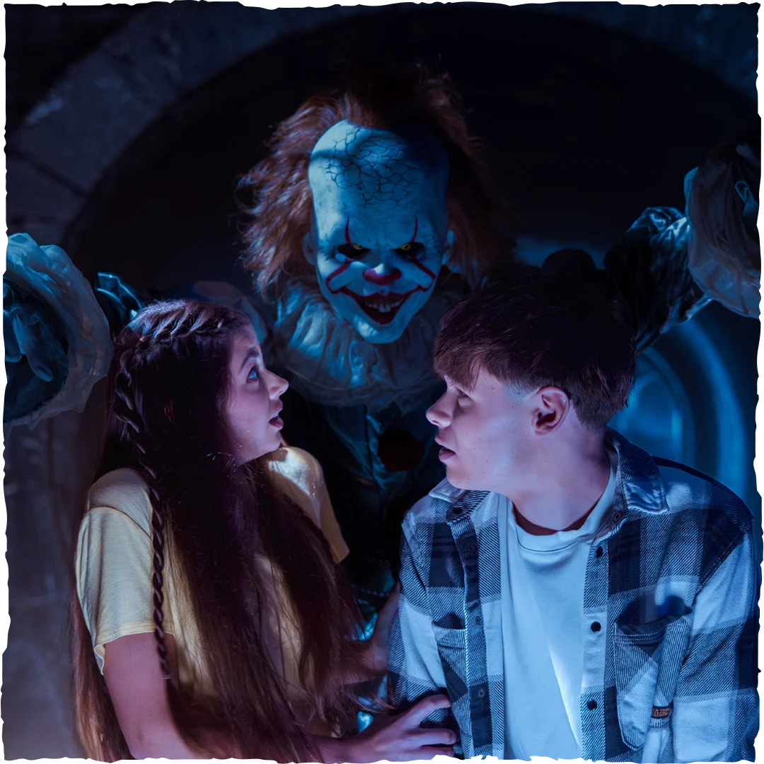 A scary clown with white face paint and red markings looms menacingly behind a frightened young woman and man, who are looking at each other in fear in a dark, eerie setting.