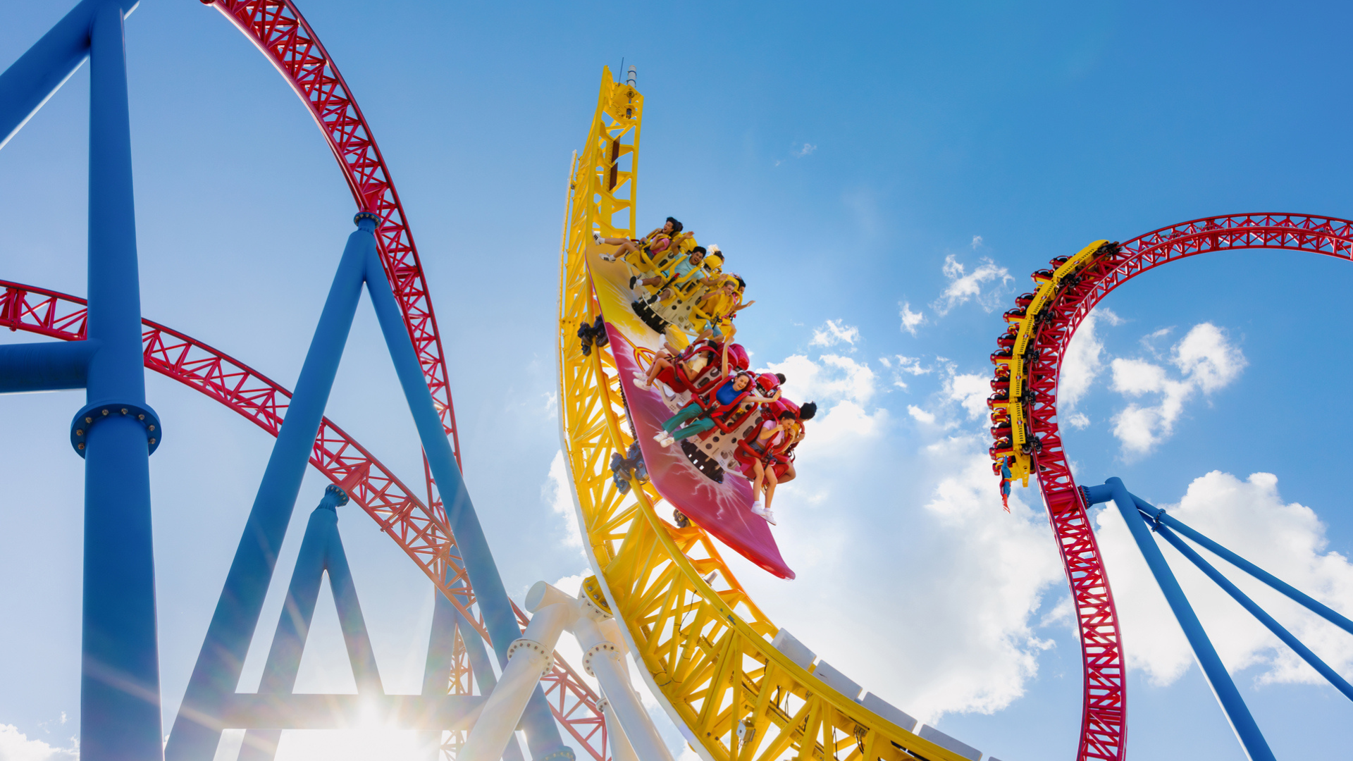 People ride a yellow and red roller coaster on a steep drop under a bright blue sky with scattered clouds and sunlight shining through the structure. The coaster features dramatic curves and loops.