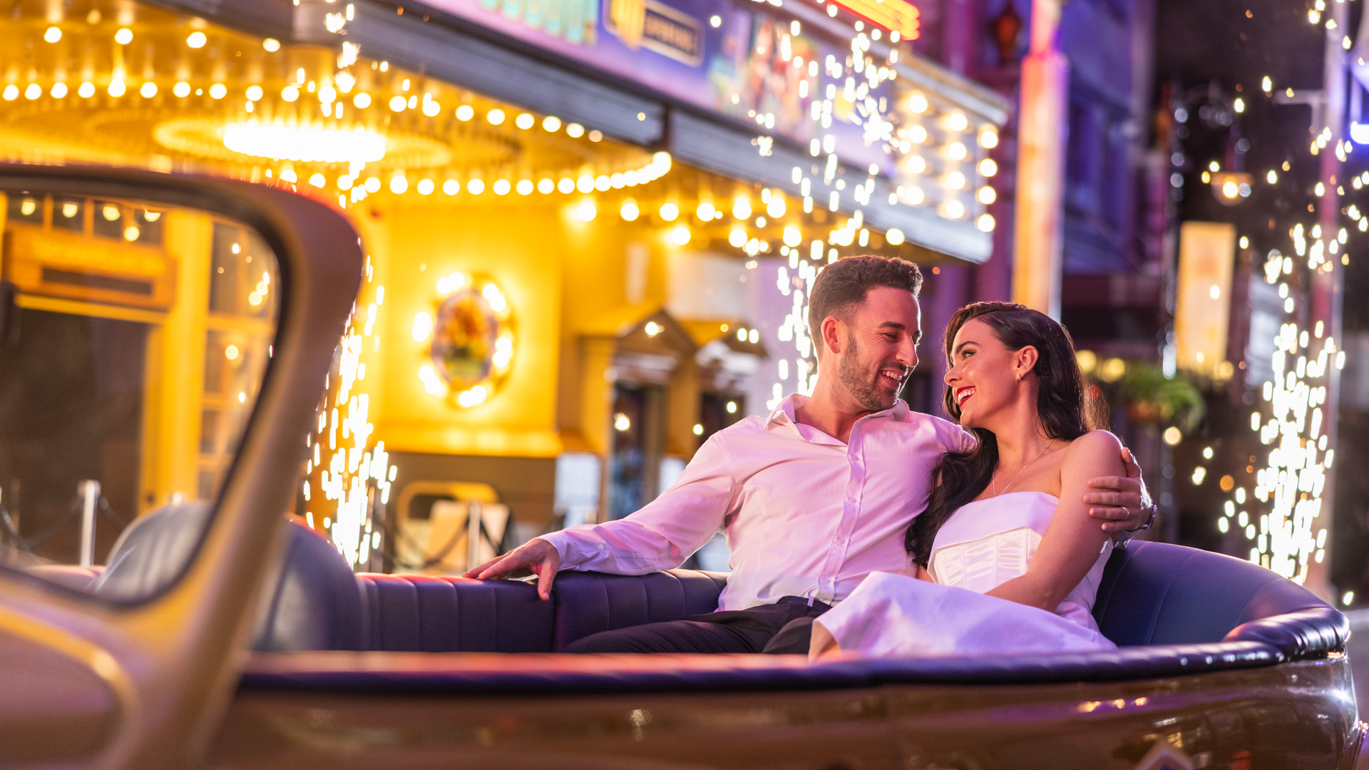 A smiling couple sits close together in a classic convertible at night, surrounded by bright lights and sparkling fireworks, with colorful buildings in the background.