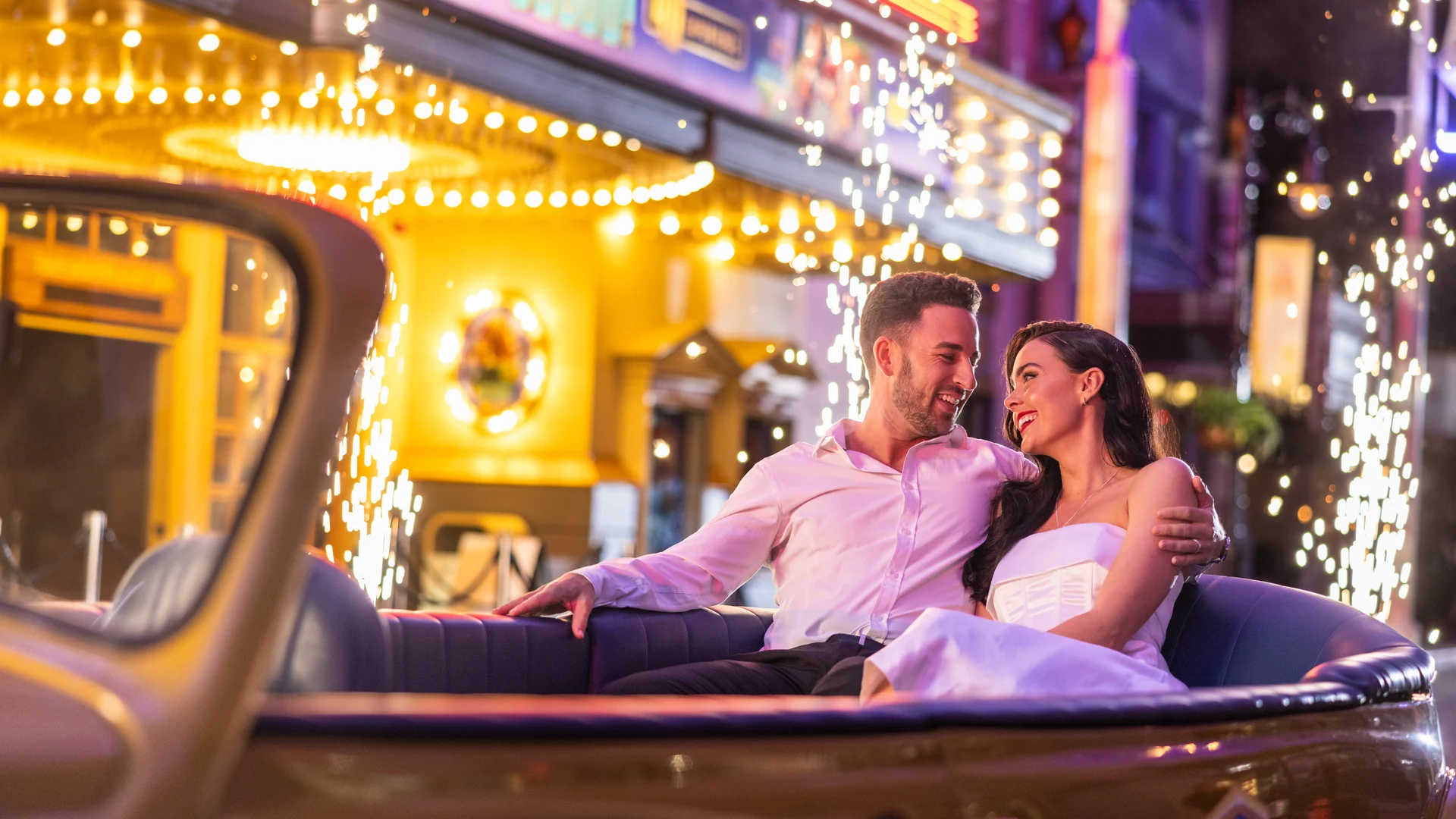 A smiling couple sits close together in a classic convertible at night, surrounded by bright lights and sparkling fireworks, with colorful buildings in the background.