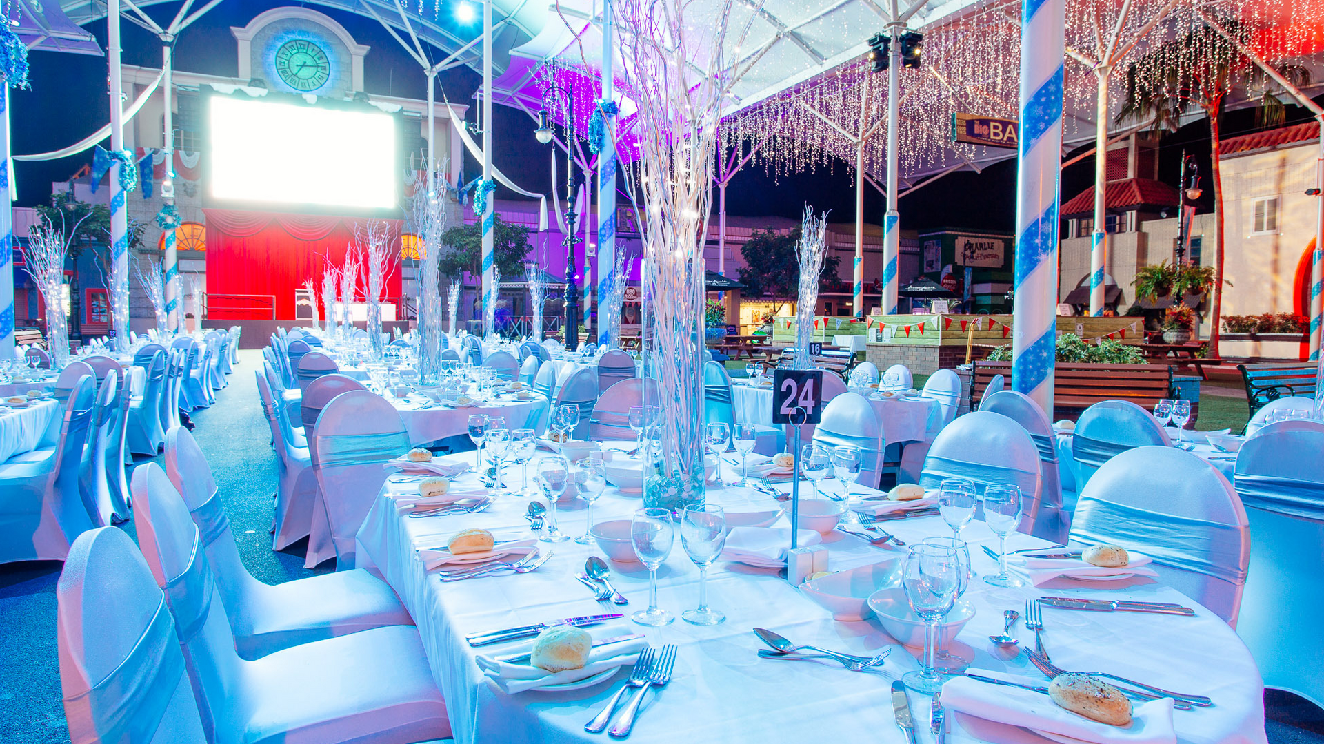 A banquet hall decorated with white and blue lighting, round tables covered in white cloths, elegant place settings, tall centerpieces, and rows of chairs, ready for a formal event or dinner party.