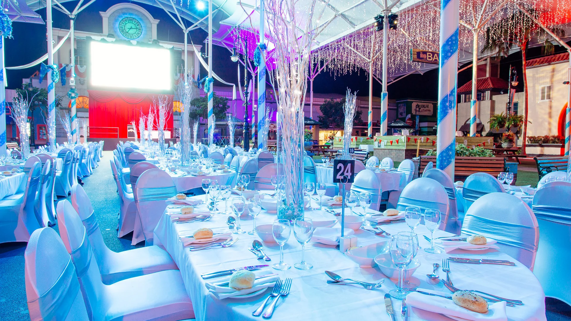 A banquet hall decorated with white and blue lighting, round tables covered in white cloths, elegant place settings, tall centerpieces, and rows of chairs, ready for a formal event or dinner party.