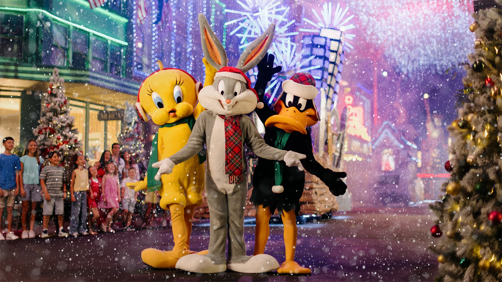 Bugs Bunny, Tweety, and Daffy Duck in festive scarves stand in front of Christmas trees and twinkling lights on a snowy street, with people watching in the background.