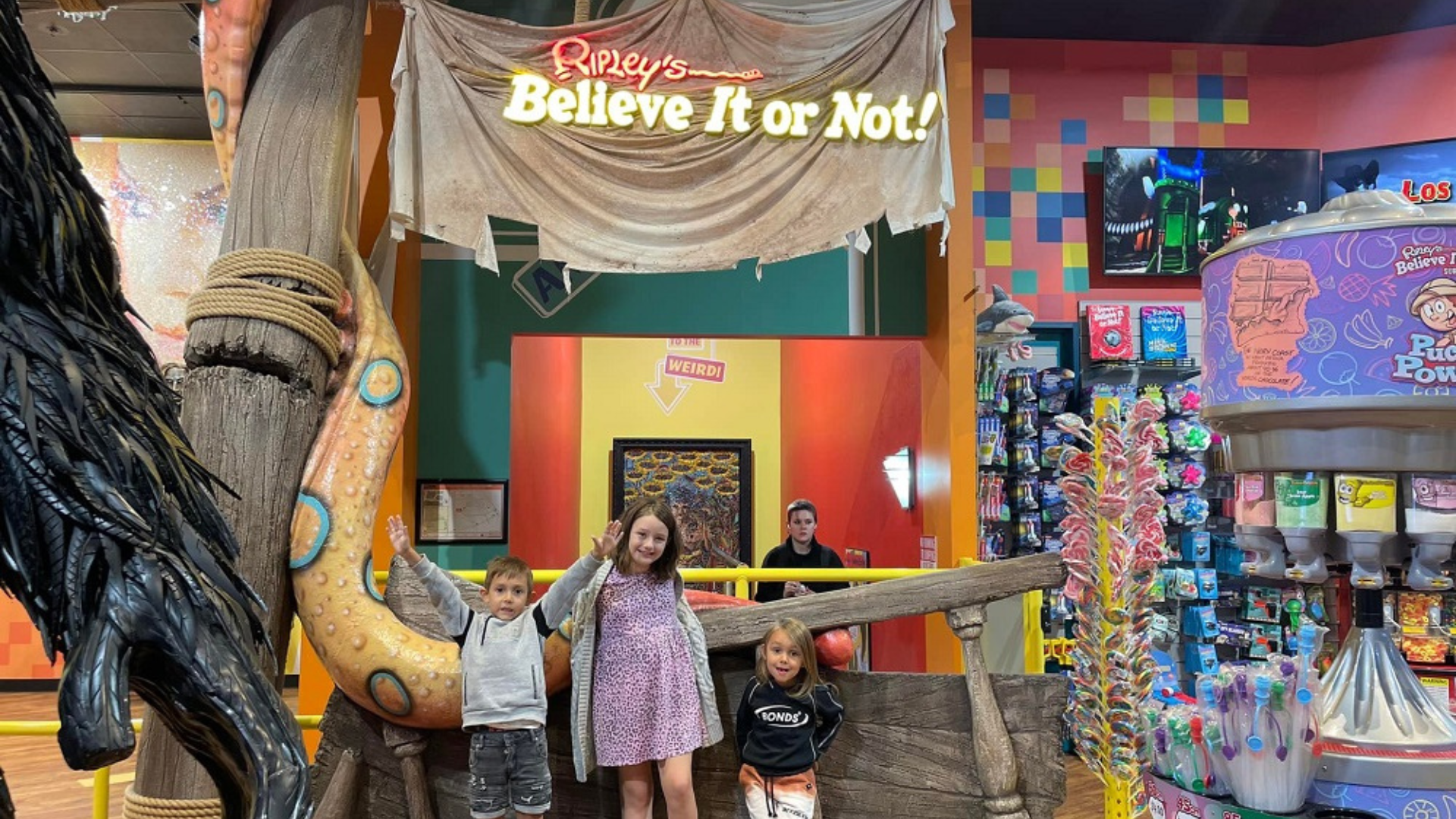 Three children stand smiling in front of a pirate ship display at Ripley’s Believe It or Not!, surrounded by colorful decor, toys, and candy displays inside a lively store.