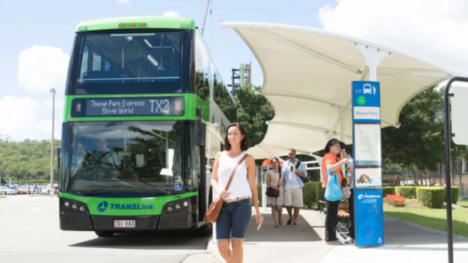 A green double-decker bus labeled "Theme Park Express" is parked at a modern outdoor bus stop. Several people, including a smiling woman in casual clothes, are walking and waiting near the bus in sunny weather.